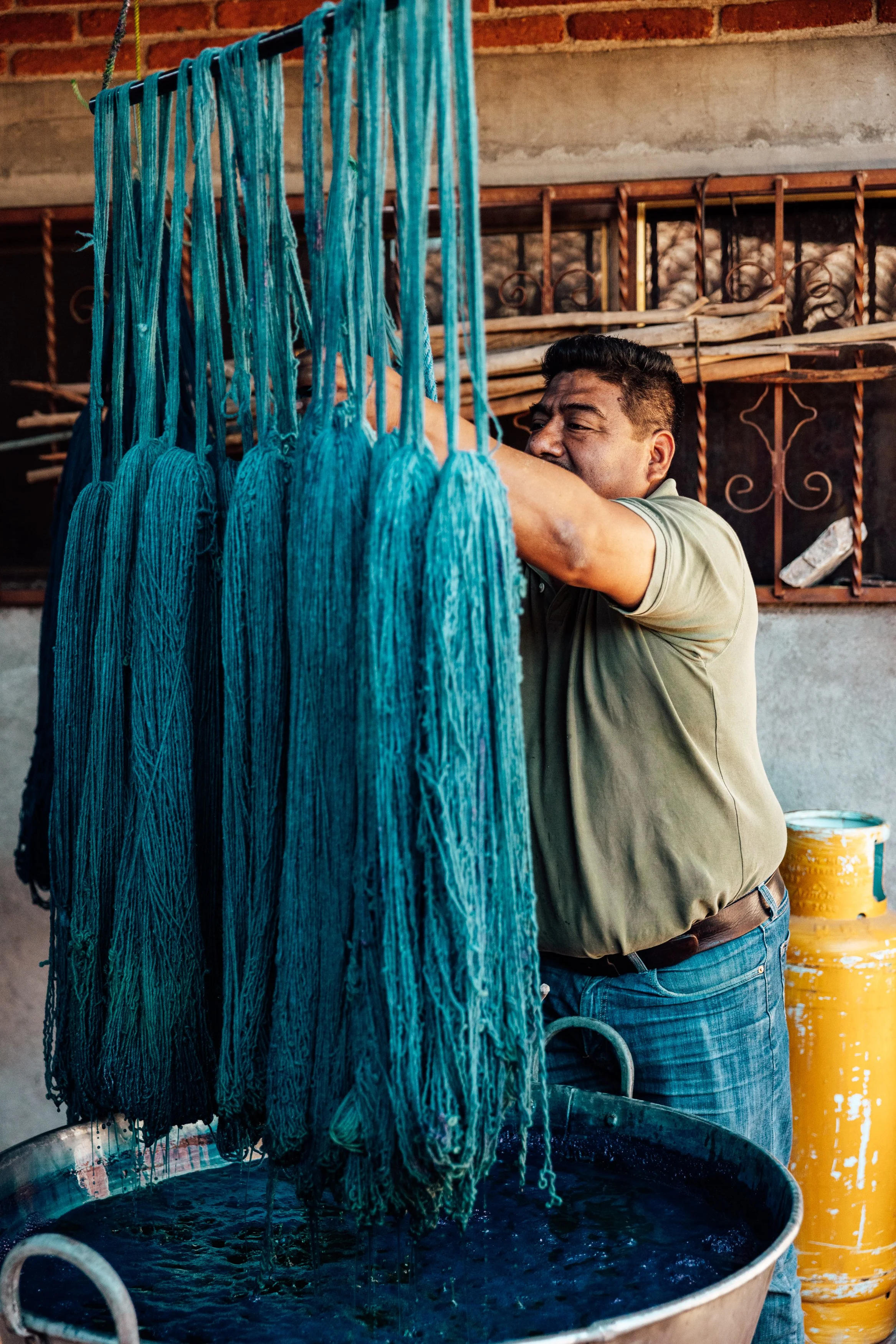 A man hanging blue yarn to dry outdoors, with a large tub of dark dye in front of him and a brick wall in the background.