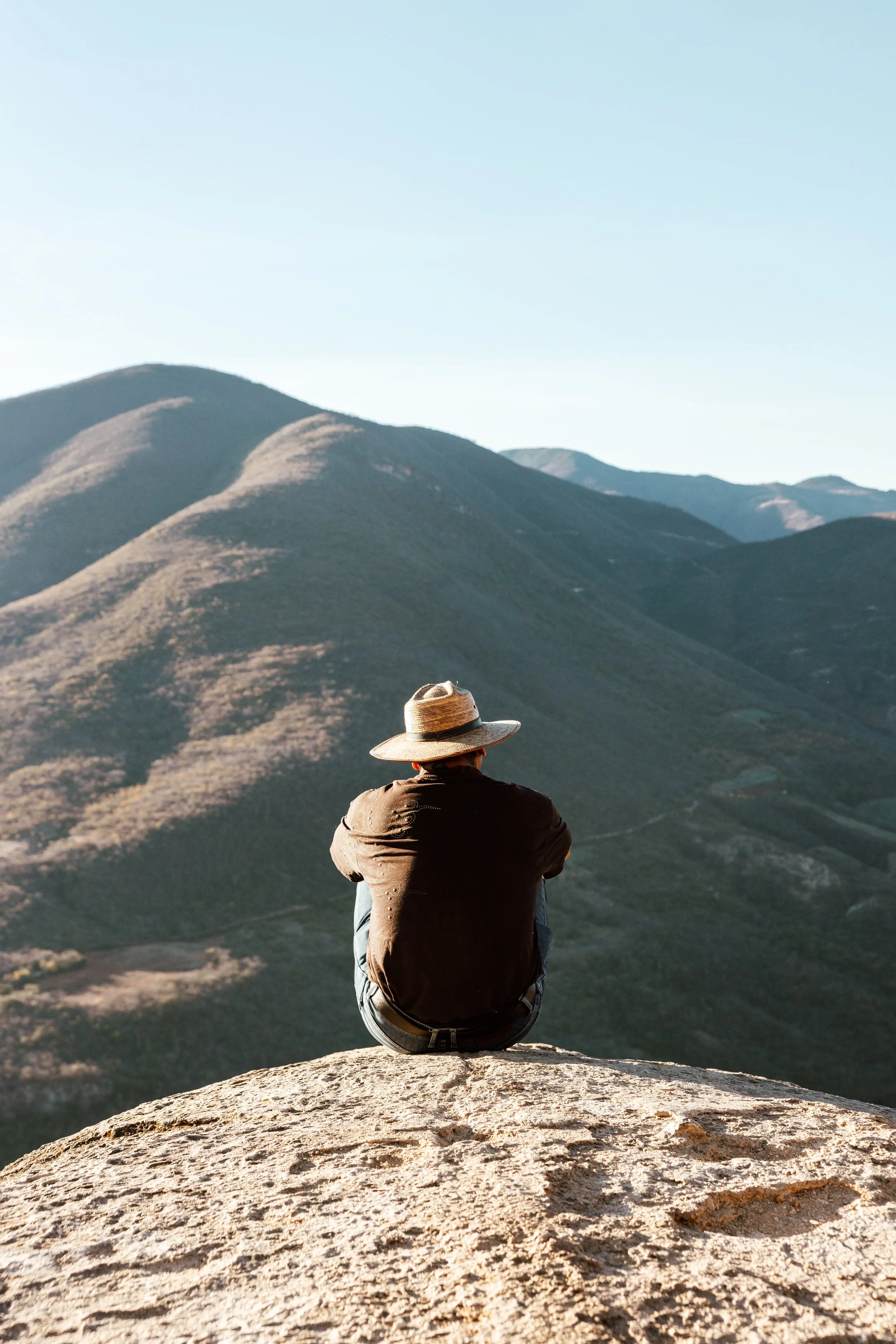 Person sitting on a large rock, facing away, wearing a hat, overlooking mountains and valleys during daylight.