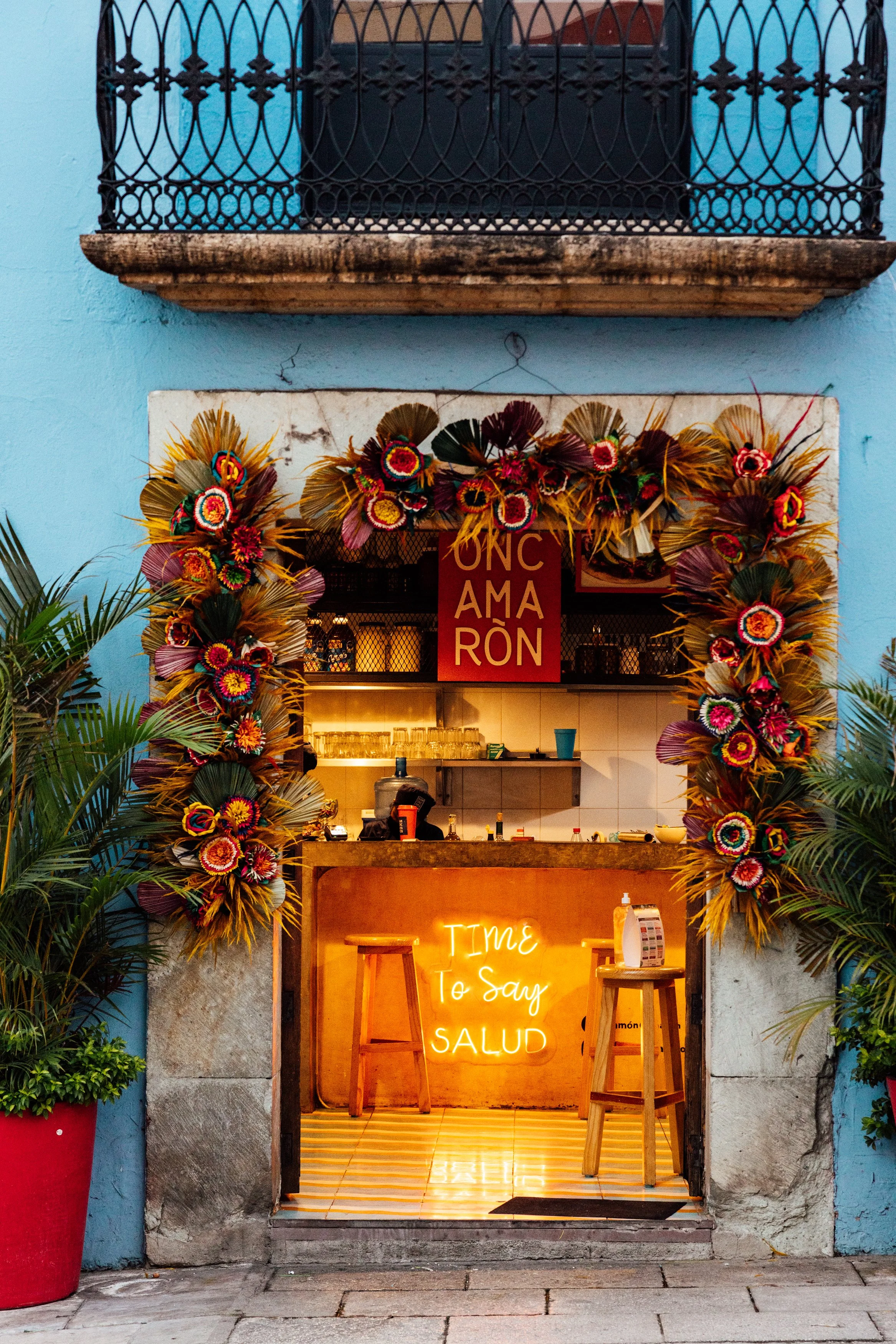Small open-air bar with a flower wreath on the entrance, neon sign that reads 'TIME TO SAY SALUD', potted plants on each side, and a sign that says 'UNC AMARÓN' above the counter, located on a city street with a blue wall.
