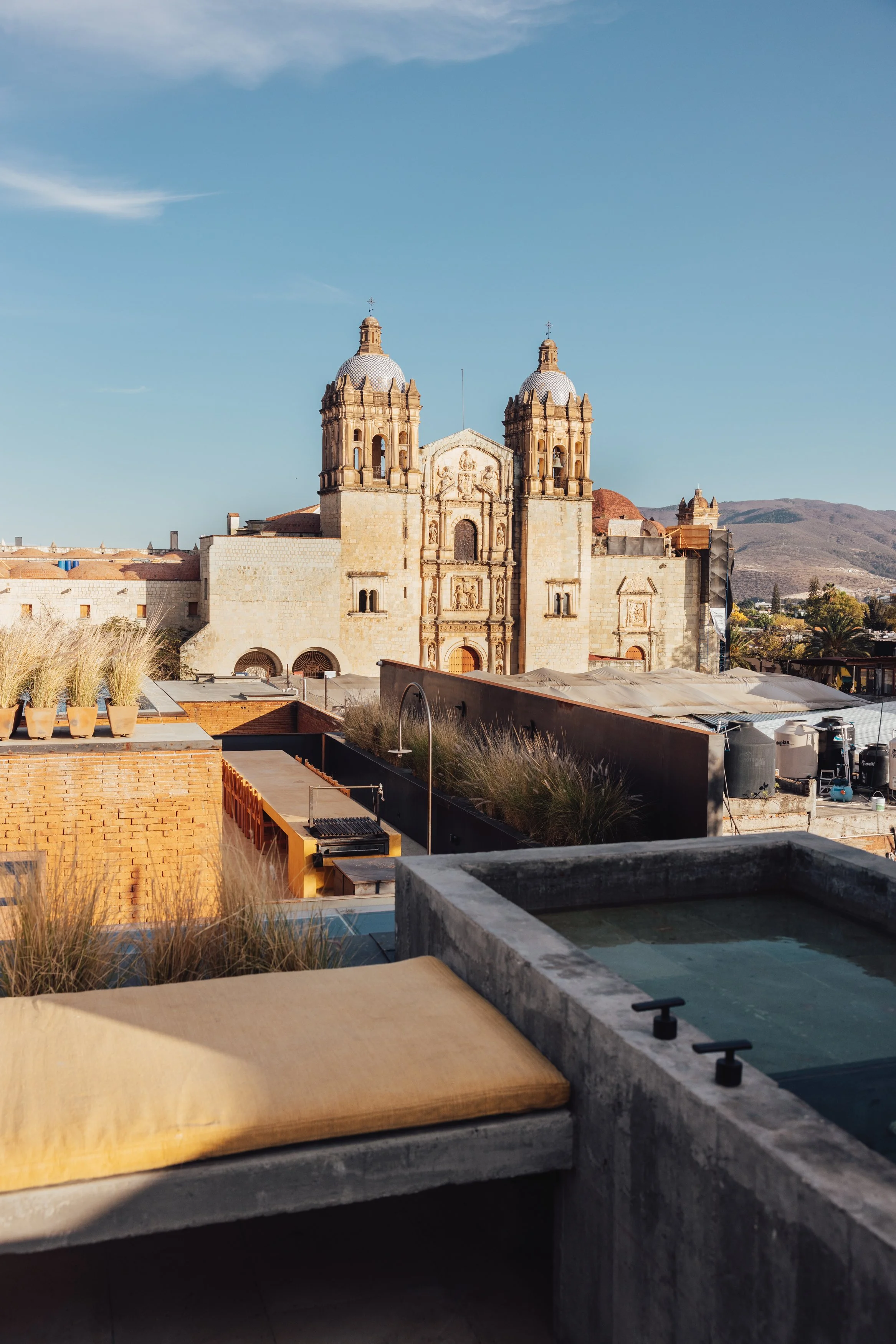 View of a historic cathedral with two bell towers, set against a mountainous landscape and blue sky, seen from a rooftop terrace with a hot tub and seating area.