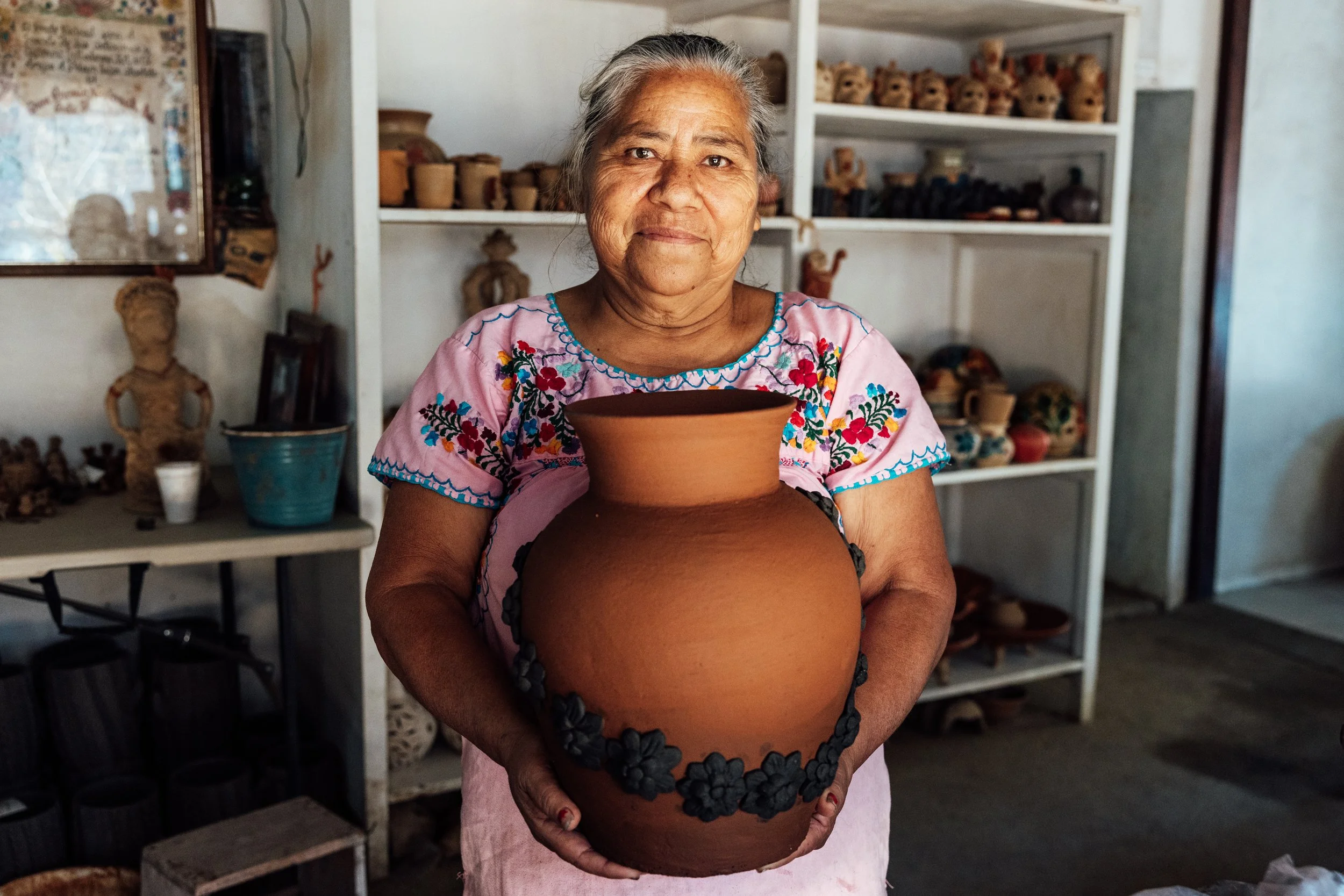 An elderly woman with gray hair, wearing a pink embroidered blouse, holding a large decorated clay pot in a pottery shop with shelves of pottery items in the background.