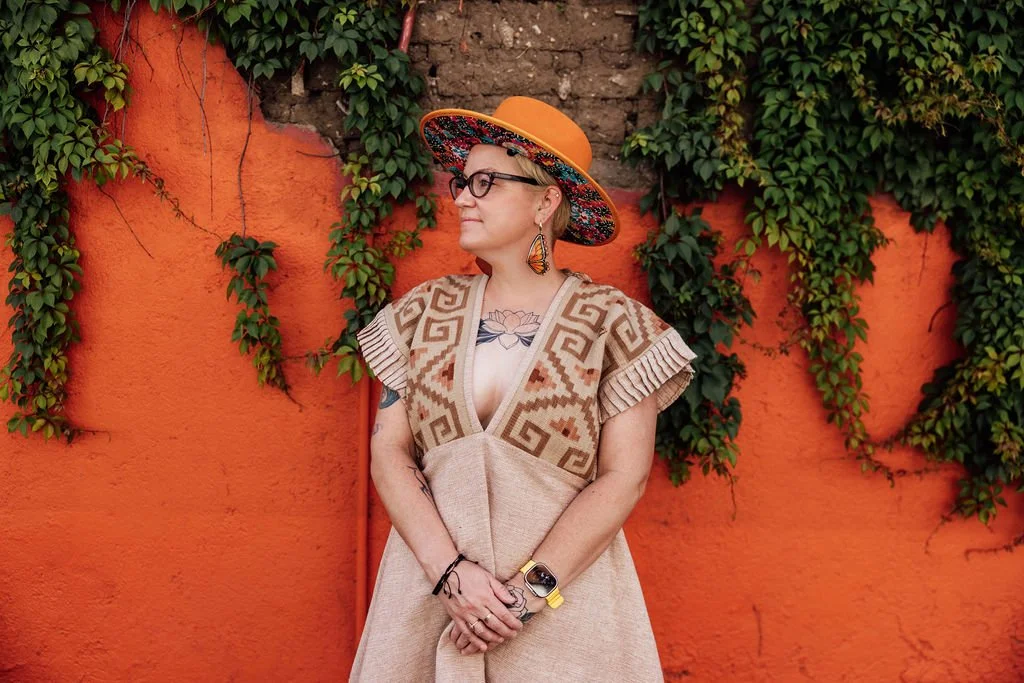 Woman with tattoos and glasses, wearing a colorful wide-brimmed hat and a patterned dress, standing in front of an orange wall covered with green vines.