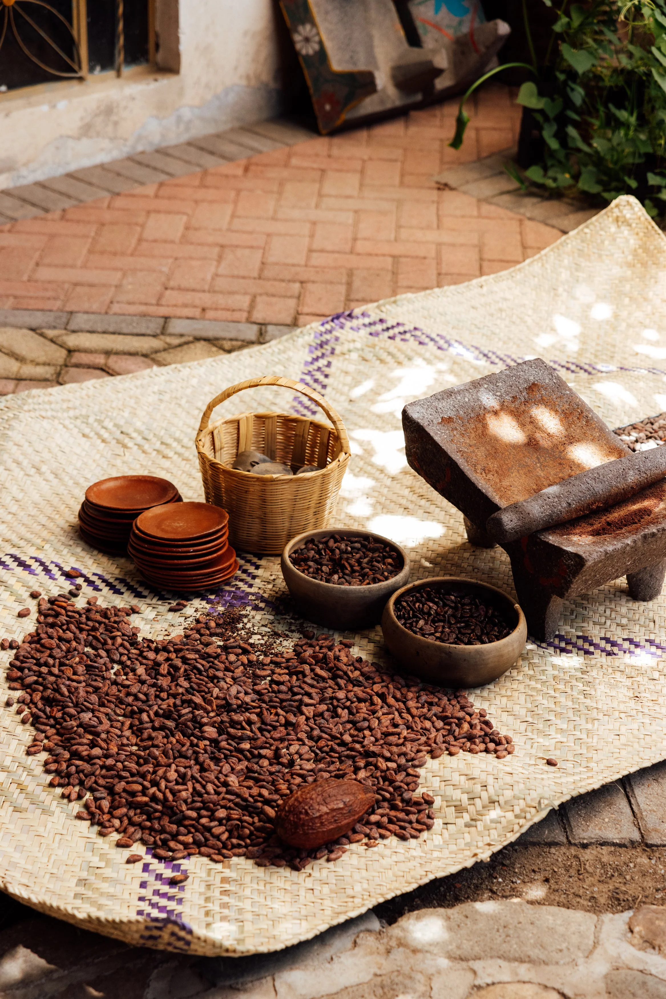 A table with roasted coffee beans, small bowls of coffee beans, a basket, an old grinder, terracotta plates, and a woven mat.