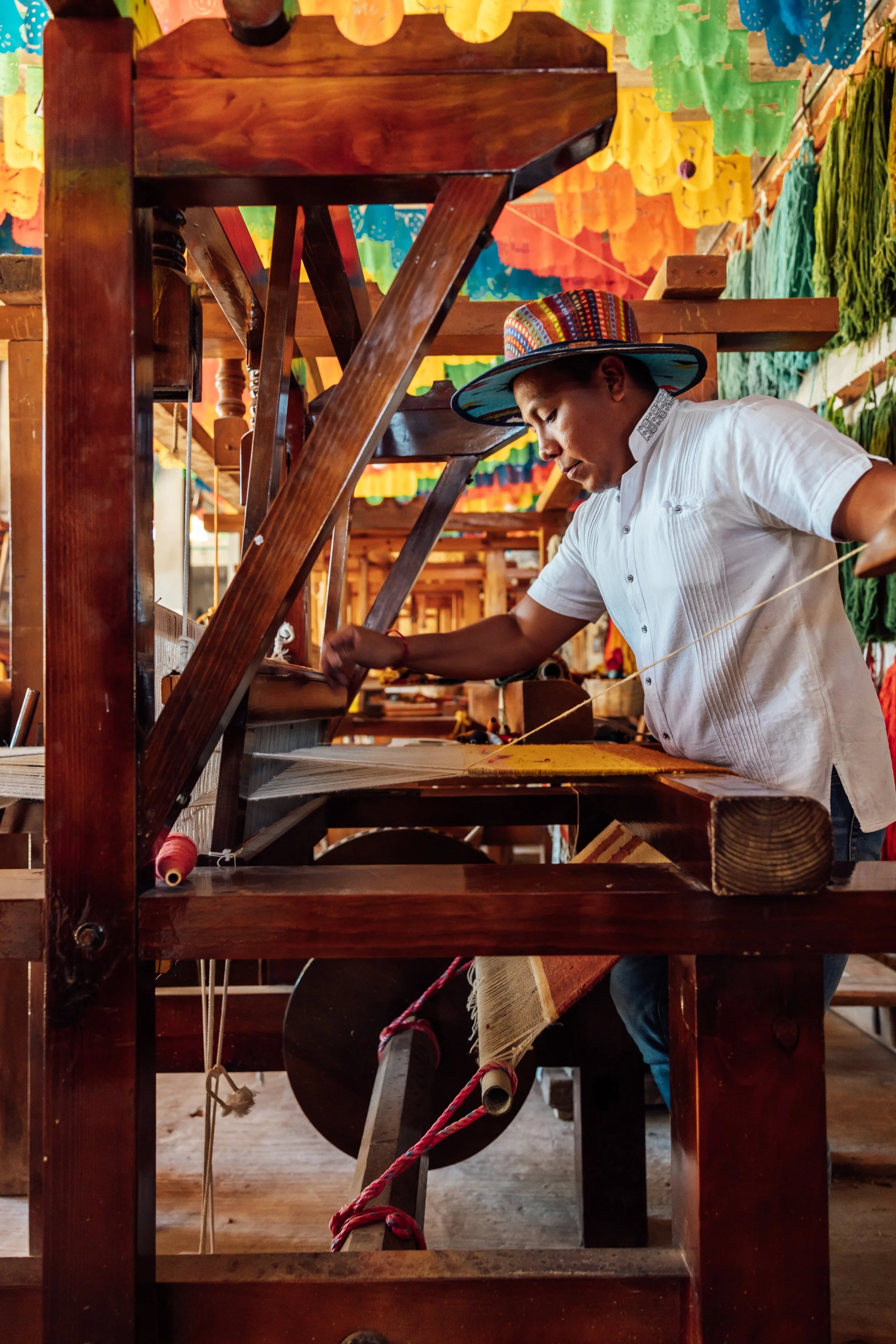 A man wearing a colorful hat and white shirt working on a traditional textile loom in a craft workshop decorated with multicolored papel picado banners.