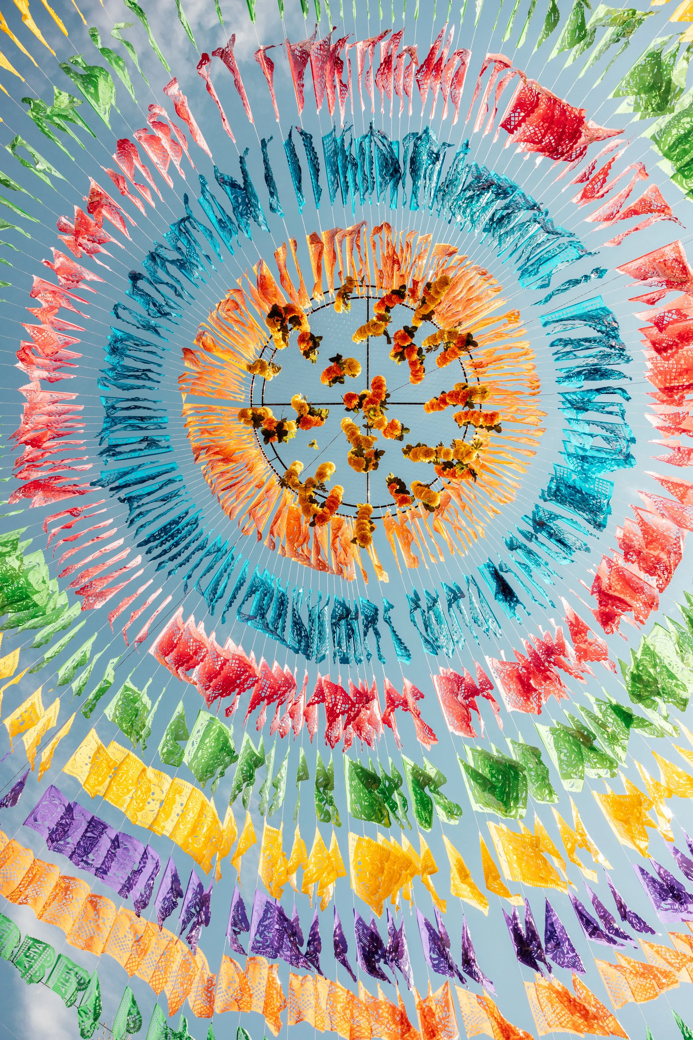 Colorful papel picado banners hanging in concentric circles against a blue sky.