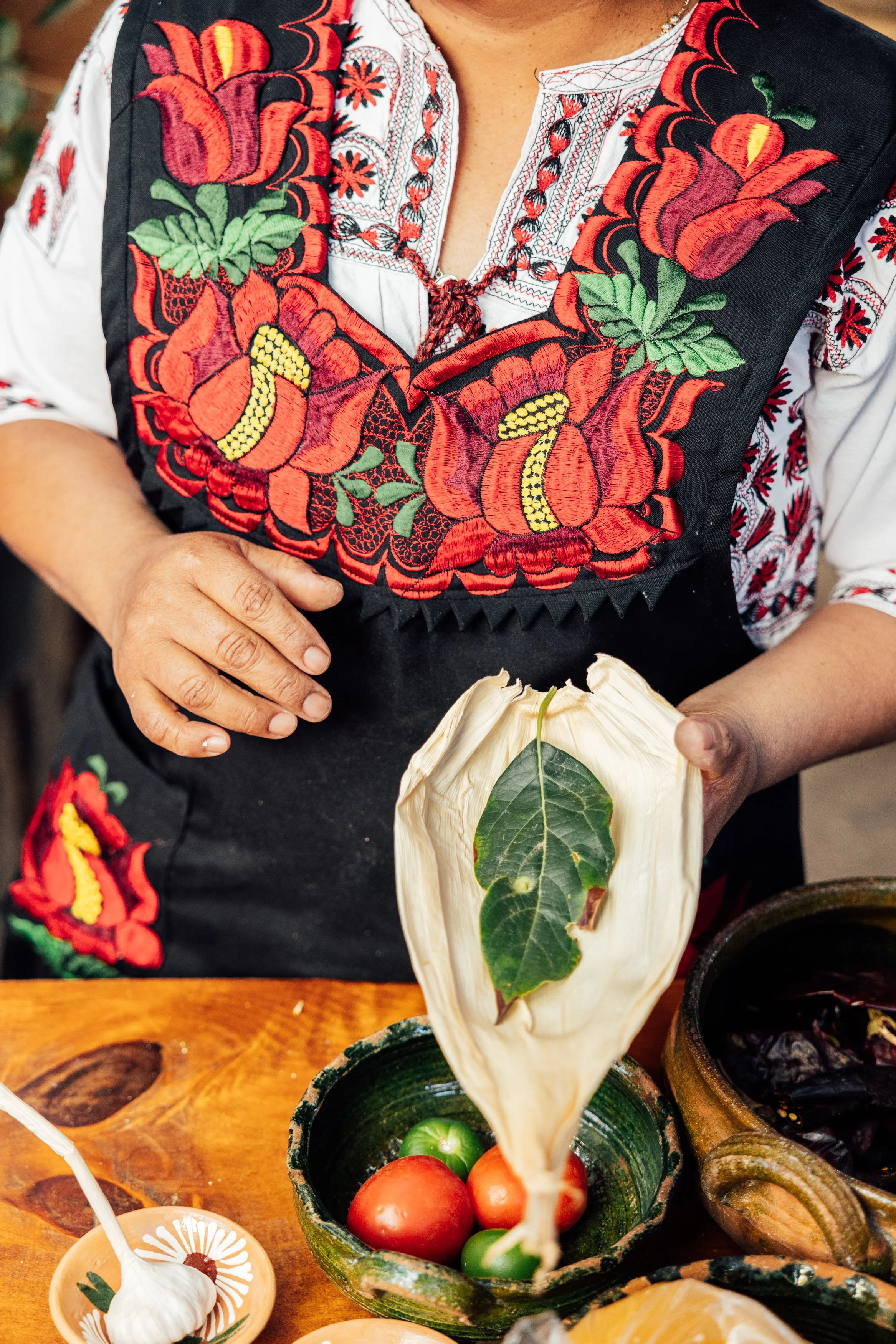 Person wearing a black apron with colorful embroidered flowers and red chili peppers, holding a bag of tomatoes and a leaf, with various herbs and vegetables on a wooden table.