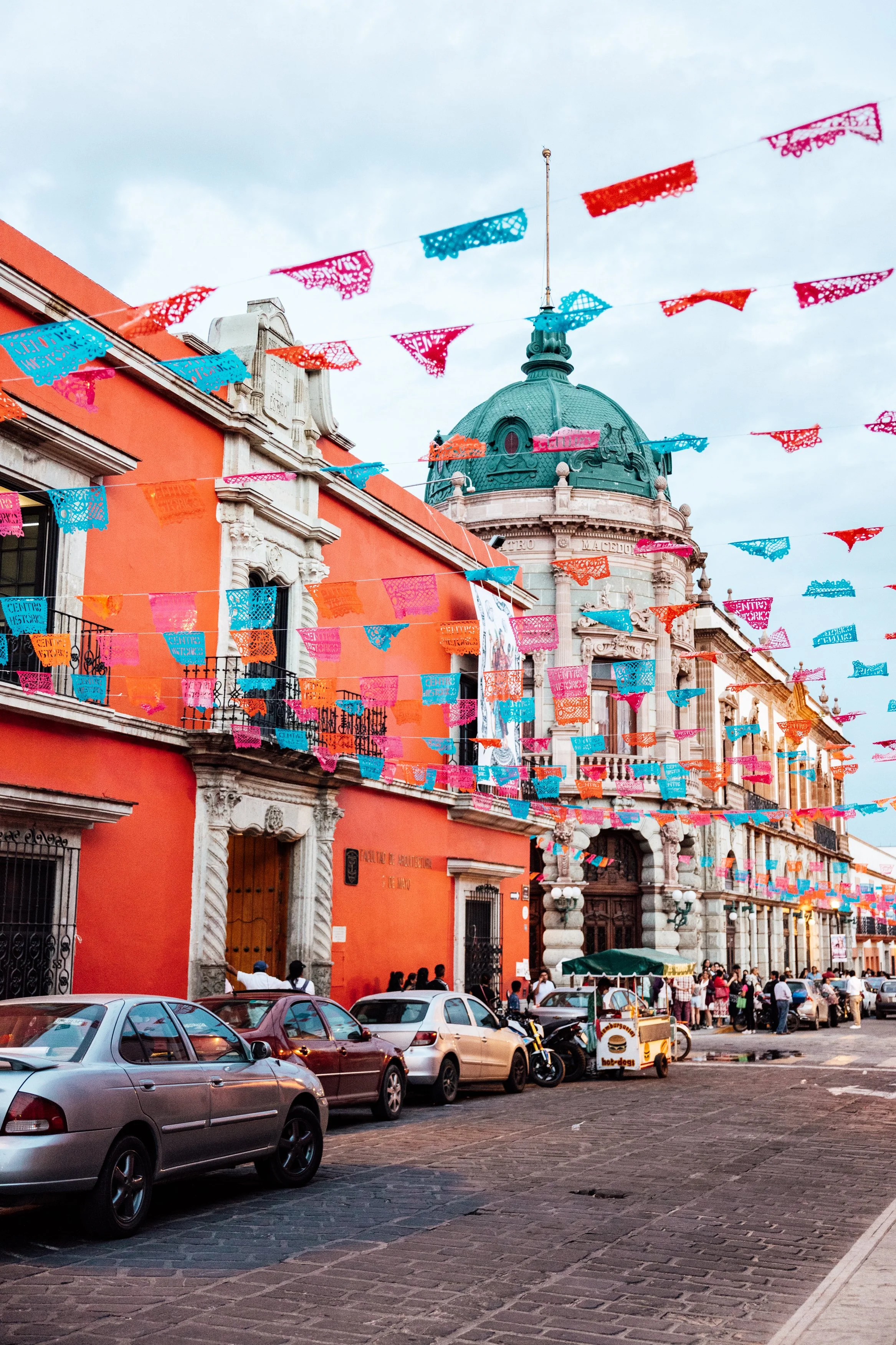 Colorful papel picado banners hanging over a historic building in a busy street, with parked cars and people walking.