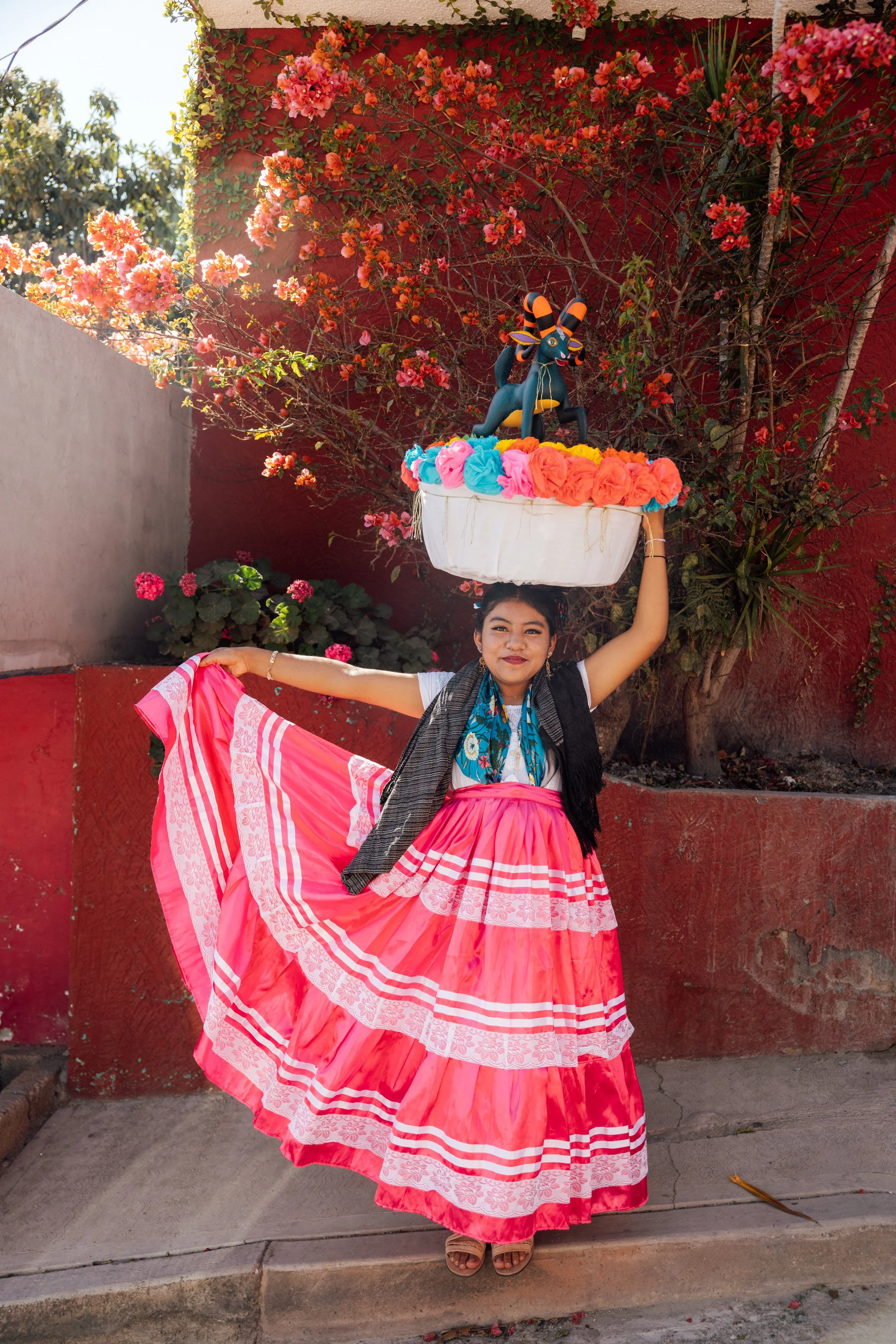 A young girl in traditional Mexican attire, wearing a long pink skirt with white and pink lace details, holds part of her skirt with one hand and balances a large basket on her head with colorful paper flowers and a small stuffed animal of a donkey."