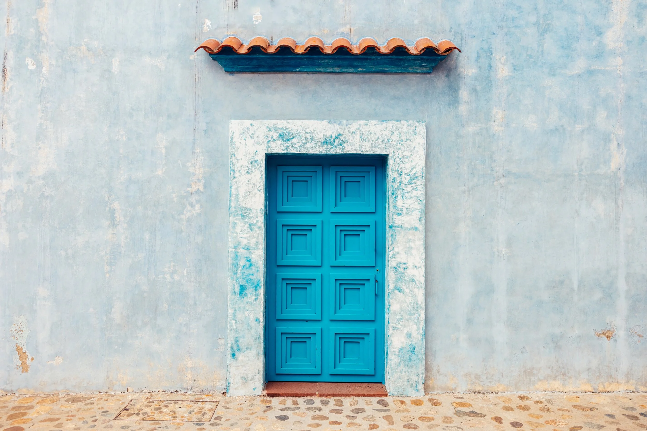 Blue door with geometric pattern set in a painted white wall with a red-tiled awning above and a stone pavement in front.