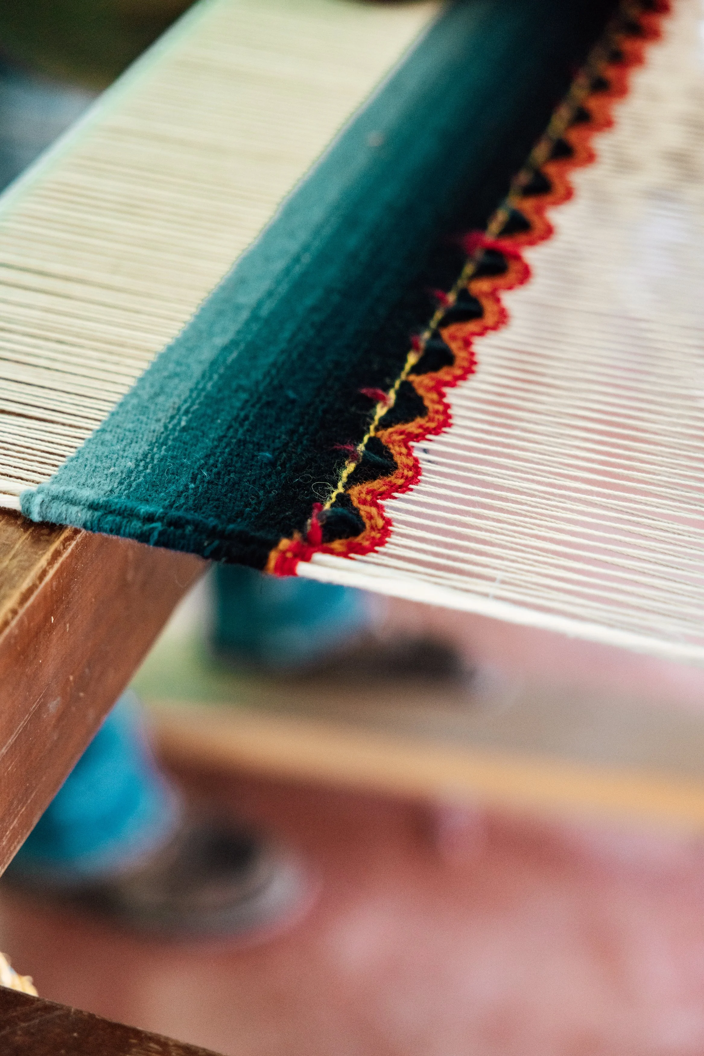Close-up of a weaving loom with partially woven fabric featuring a decorative border with red, yellow, and black zigzag pattern.