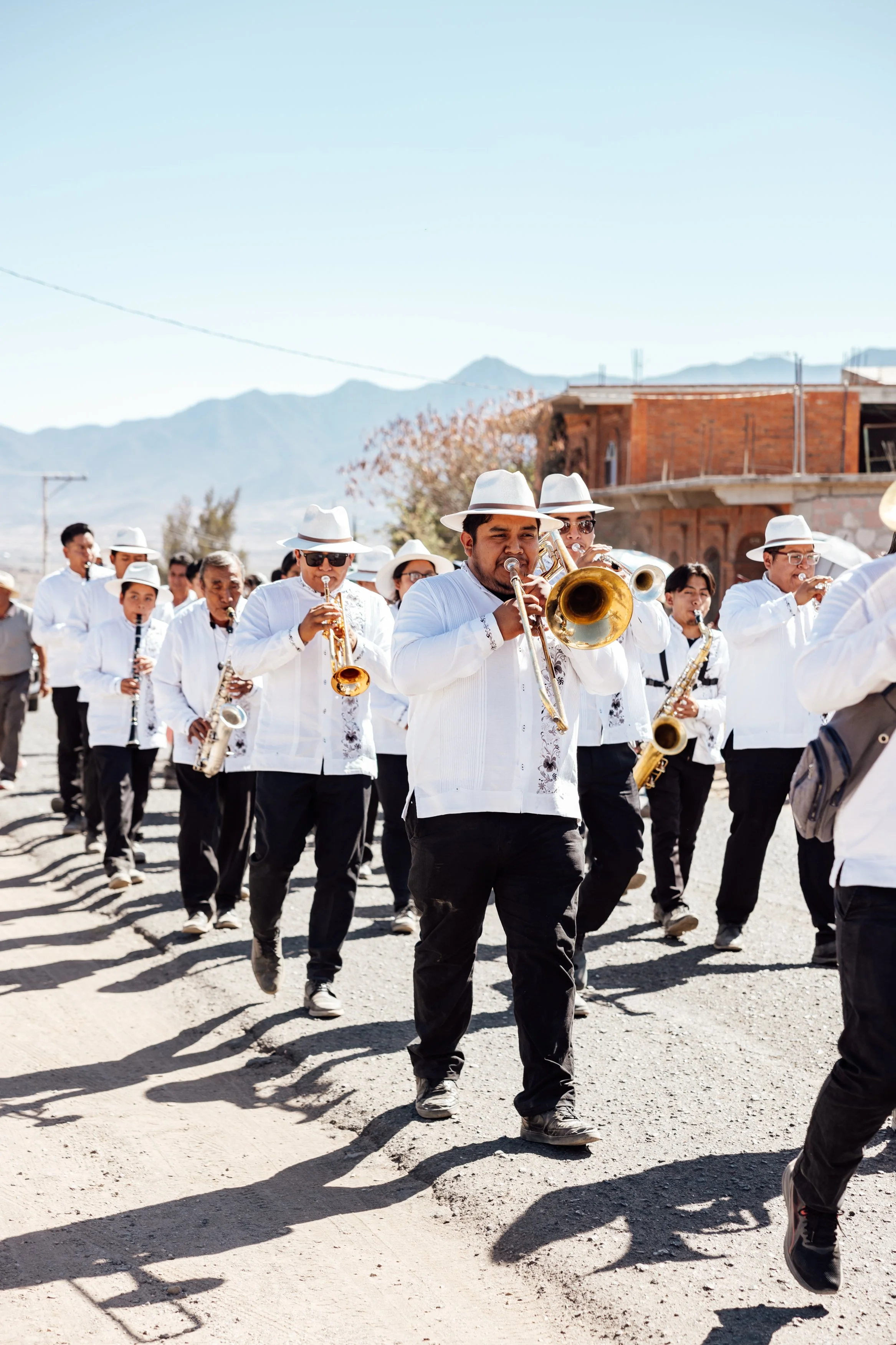 A marching band performing outdoors, dressed in white shirts, black pants, and white hats, with mountains in the background.