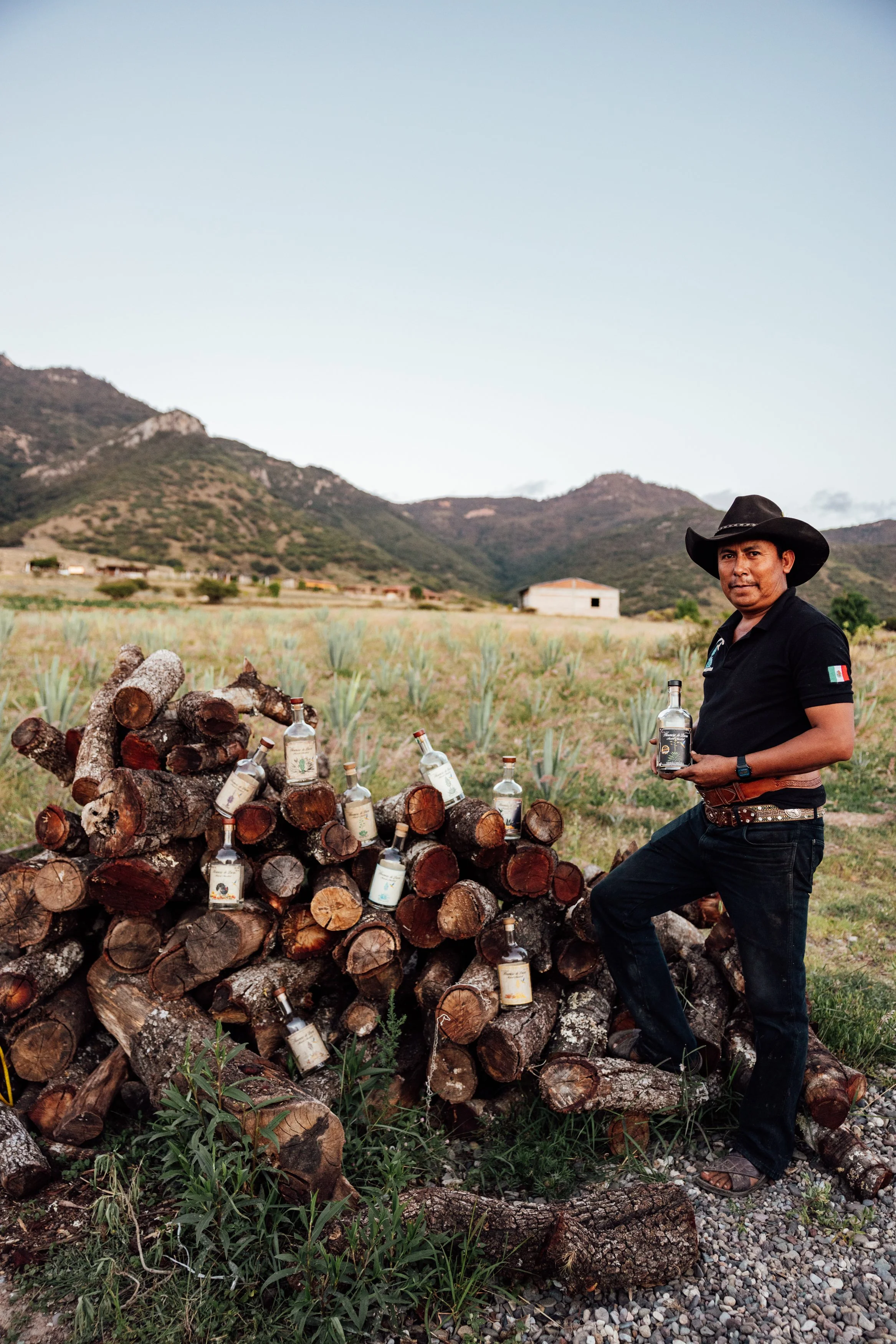 A man wearing a black cowboy hat and black shirt stands next to a large pile of chopped firewood logs with bottles of tequila placed on top of the logs. He is holding a bottle of tequila in his right hand, outdoors in a rural landscape with mountains