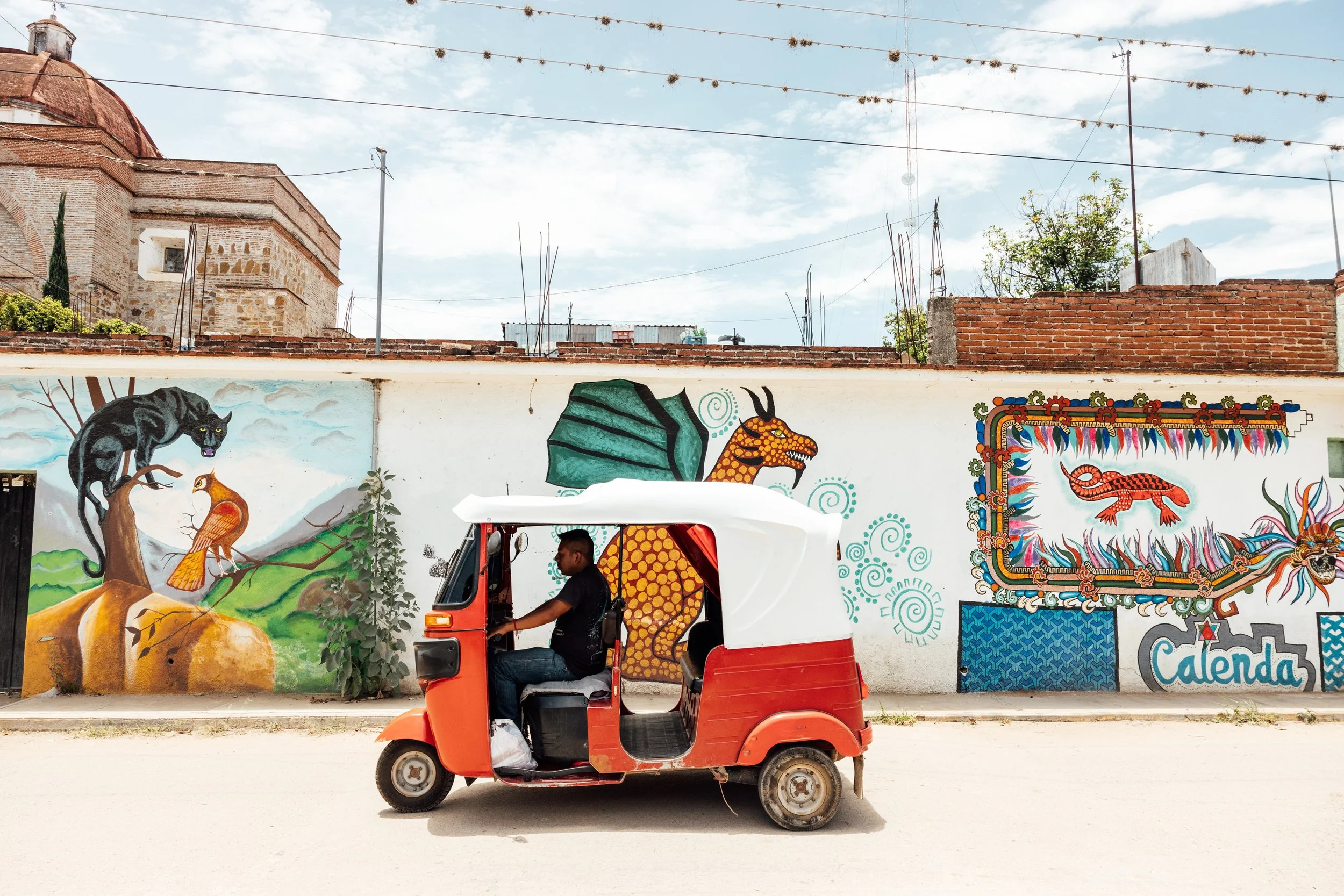 A man in a black shirt riding a red and white tuk-tuk in front of a colorful mural featuring a black panther, a bird, a dragon, and abstract designs, with a wall labeled 'Calenda' on the right.