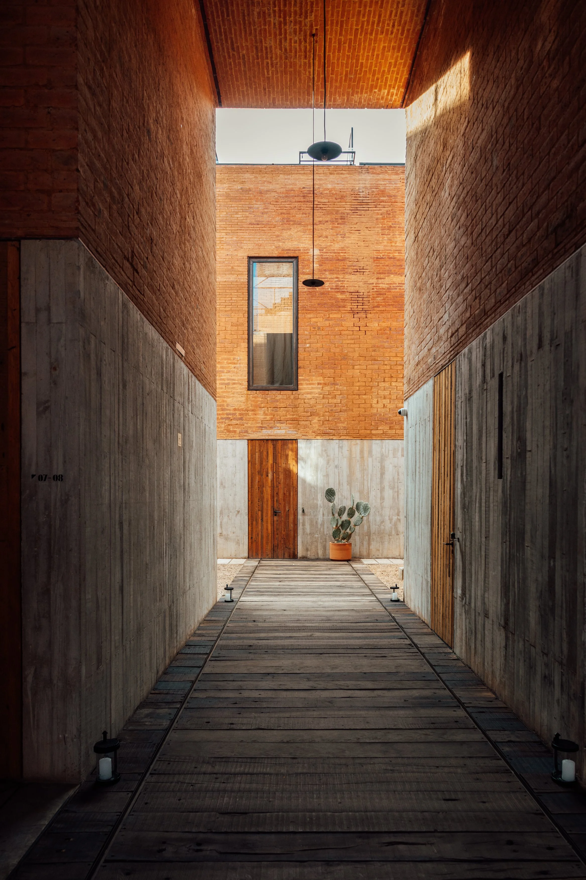 Indoor corridor with brick and concrete walls, wooden floor, potted cactus plant, wooden door, and at the end of the corridor, a concrete wall with wooden door and a window.