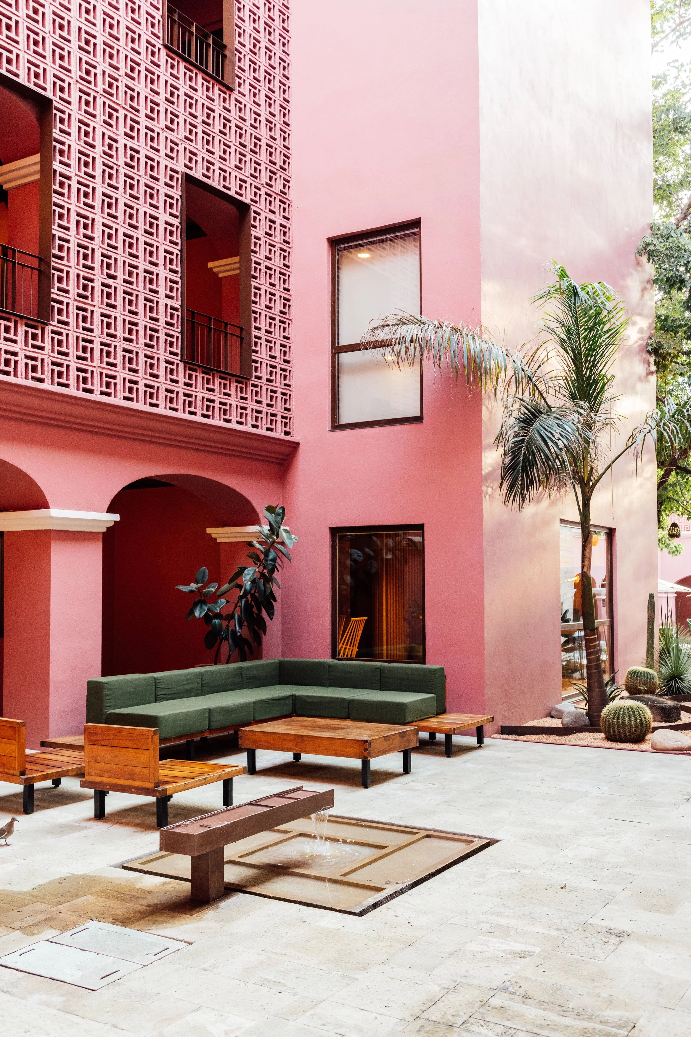 A pink building with decorative latticework and arched entryways, a courtyard with green outdoor seating, a water fountain, and desert plants.