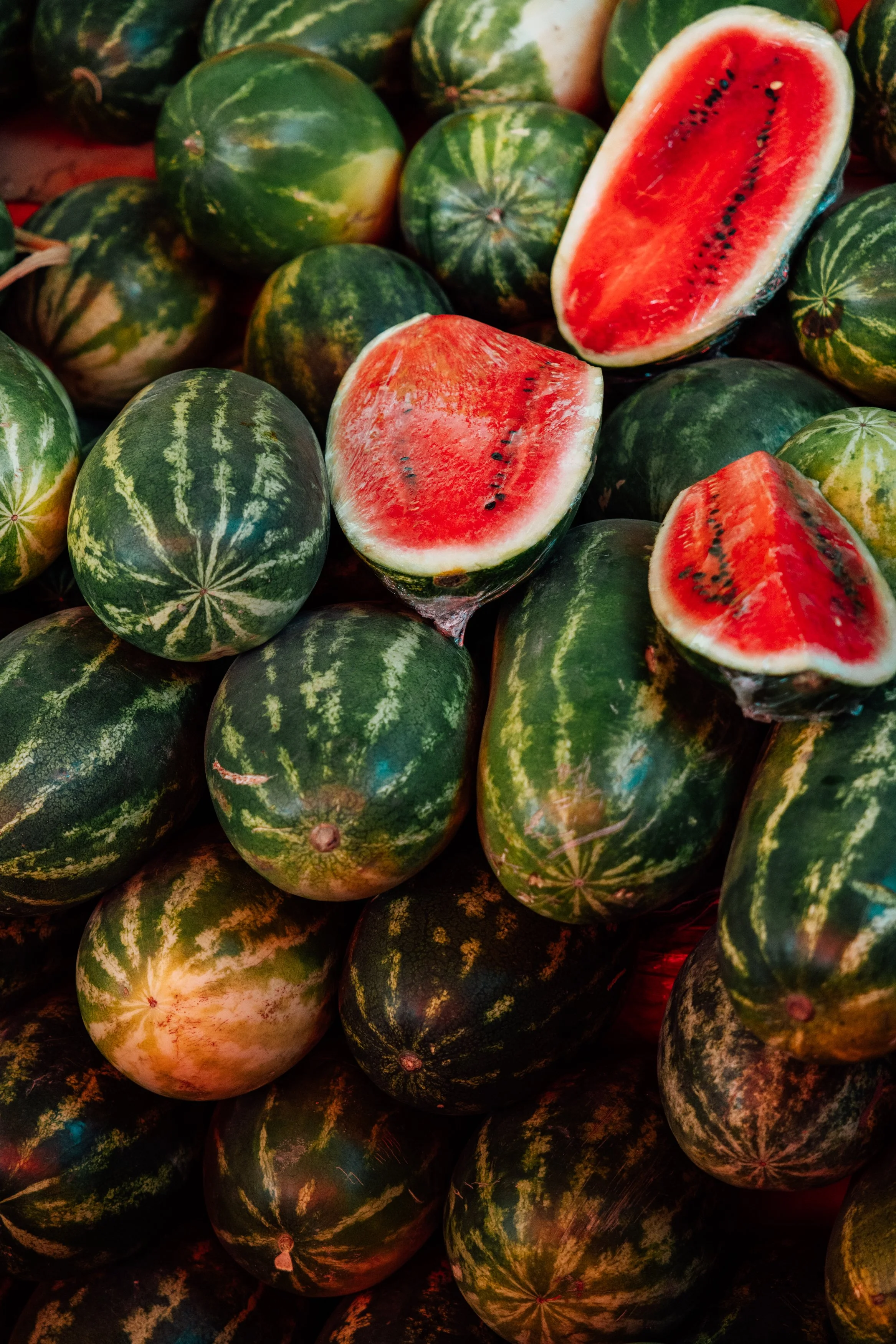 Several green watermelons with dark green stripes, some cut open to show red flesh and black seeds, piled together.
