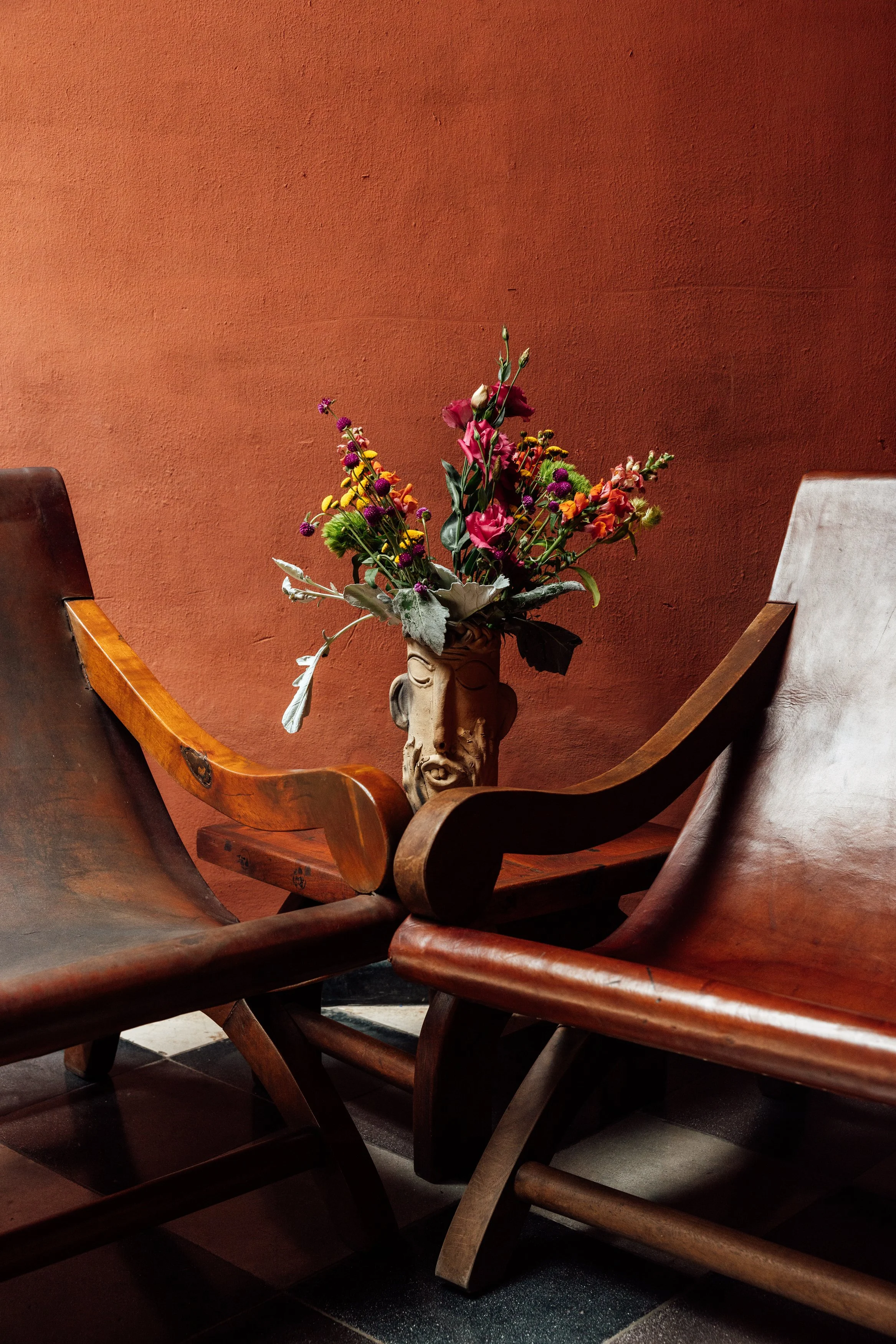 A bouquet of colorful flowers in a decorative ceramic vase, placed between two wooden chairs against a reddish-orange wall.
