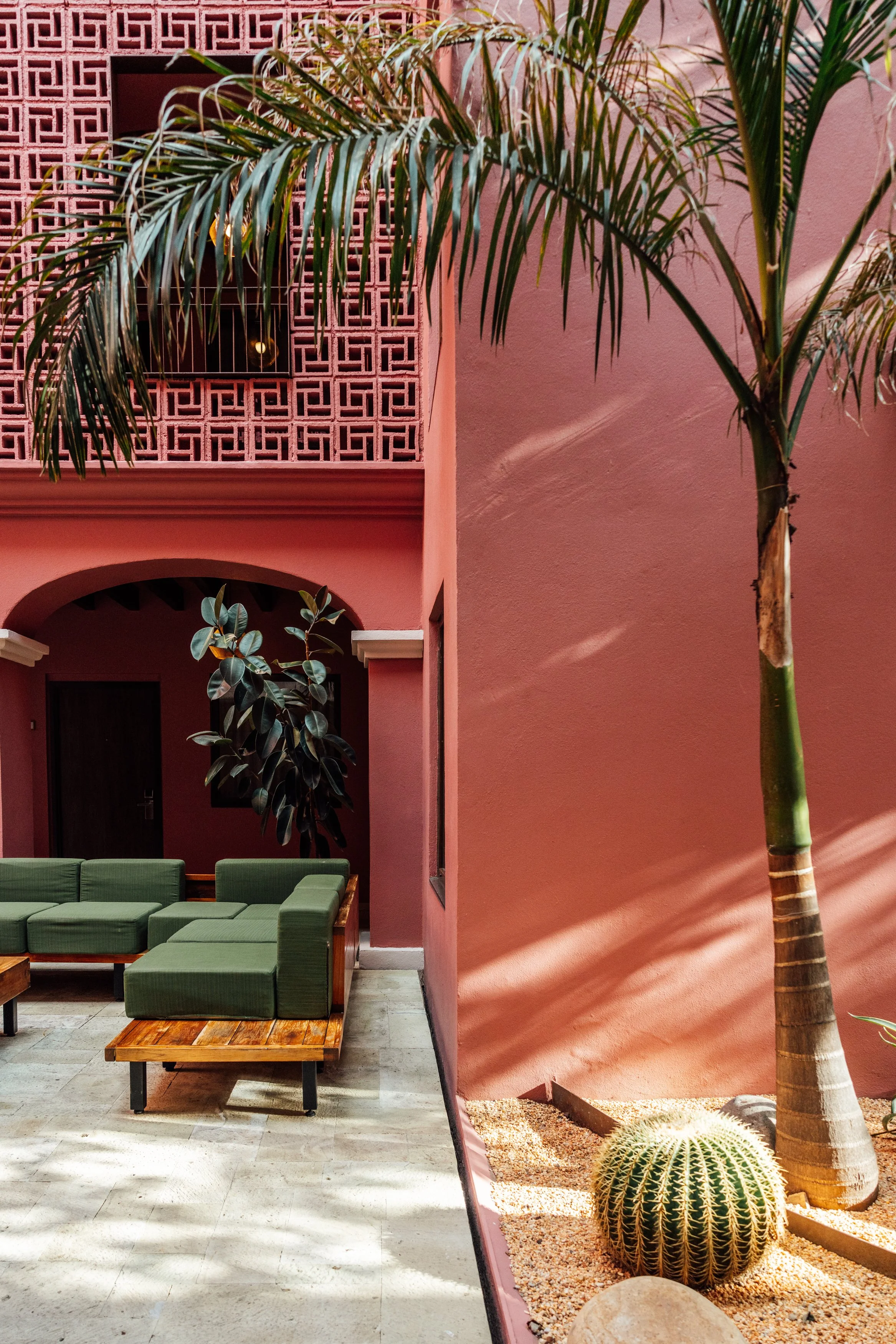 A courtyard with pink walls, a green sofa, palm trees, and a spherical cactus on gravel.