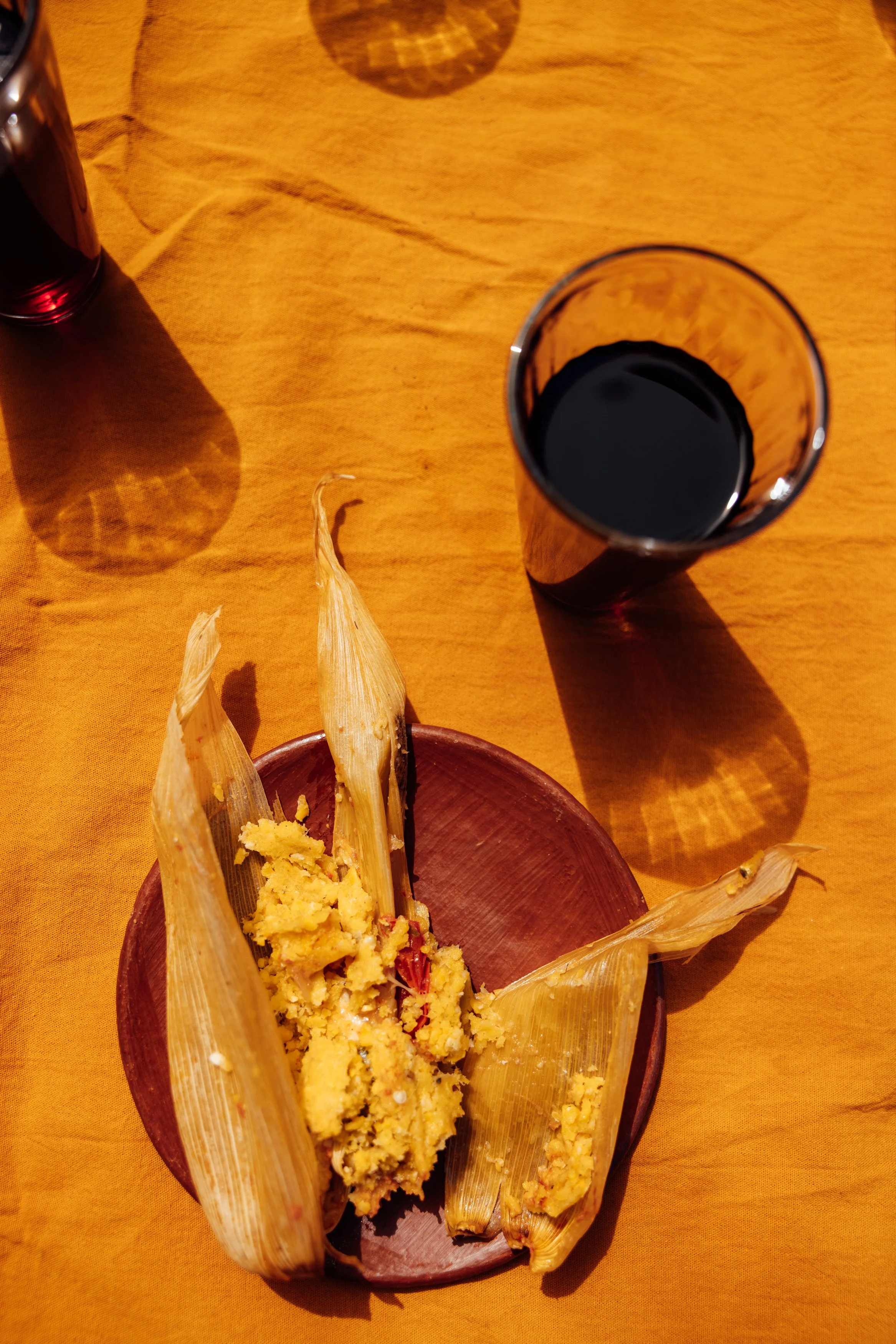 A plate with tamales, some partially unwrapped, on a yellow tablecloth next to a glass of dark soda.