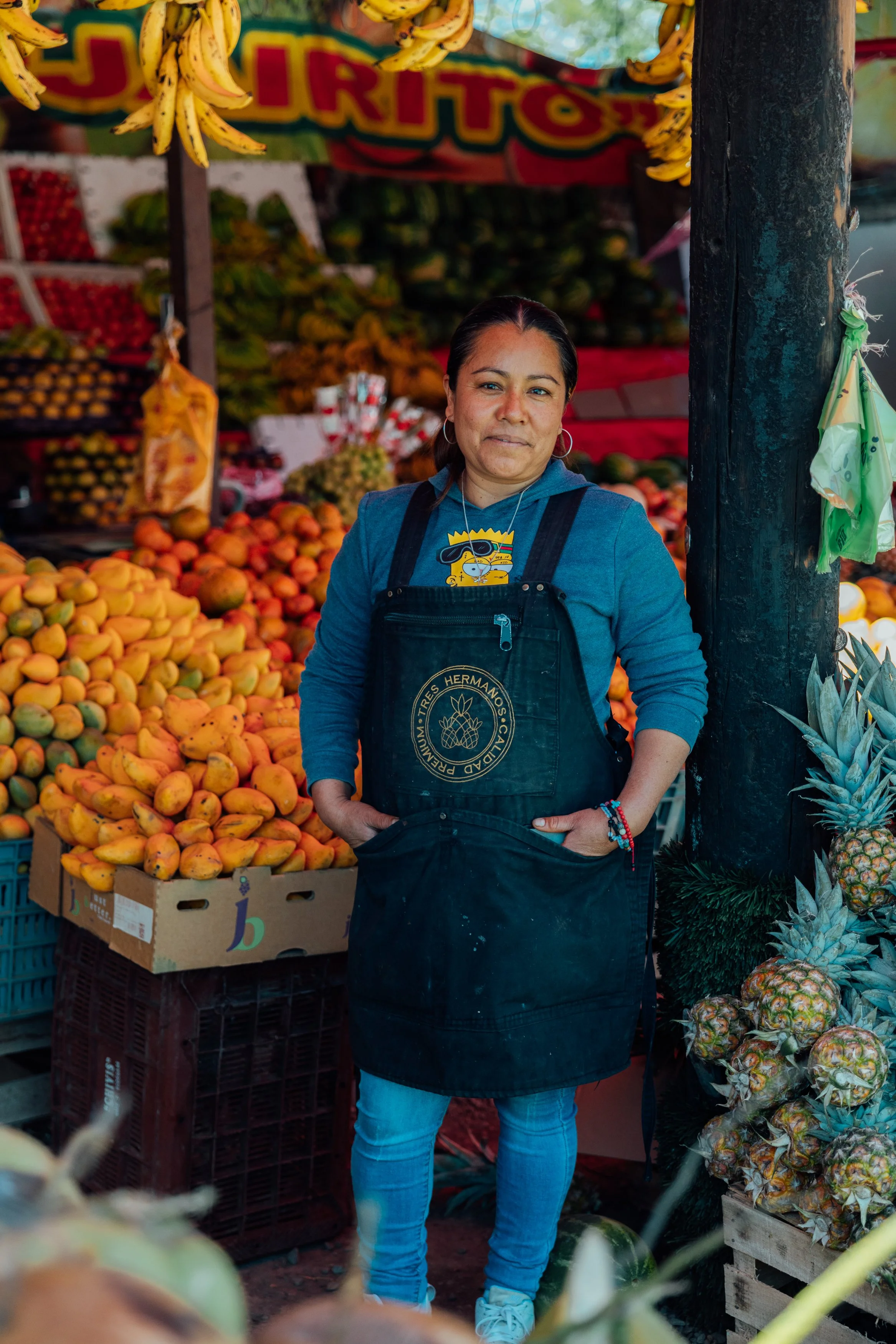 A woman standing behind a fruit stand at an outdoor market, with bananas, mangoes, pineapples, and other fruits displayed around her.
