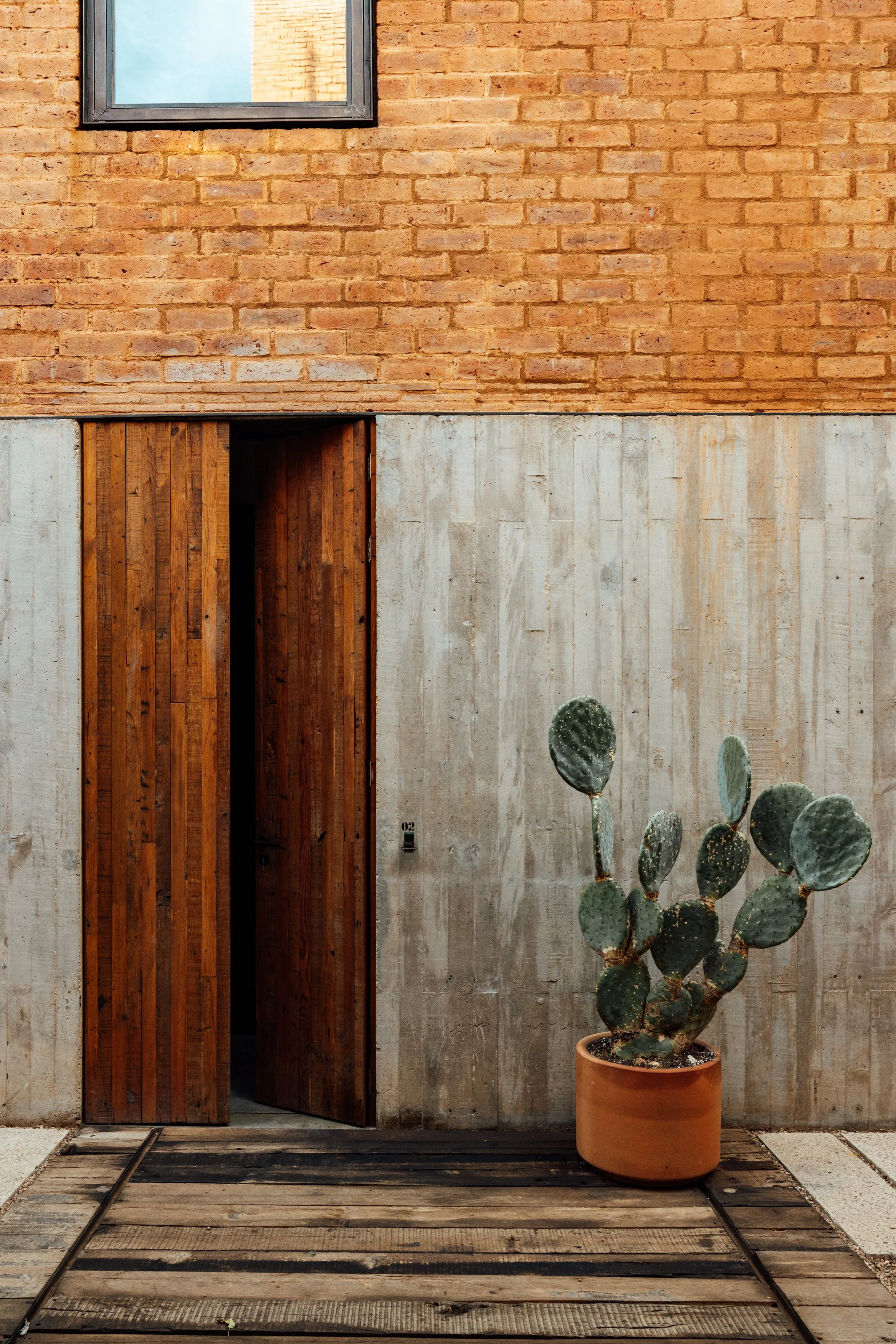 A modern building exterior with a brick upper wall, concrete lower wall, wooden door, and a cactus in a terracotta pot outside.