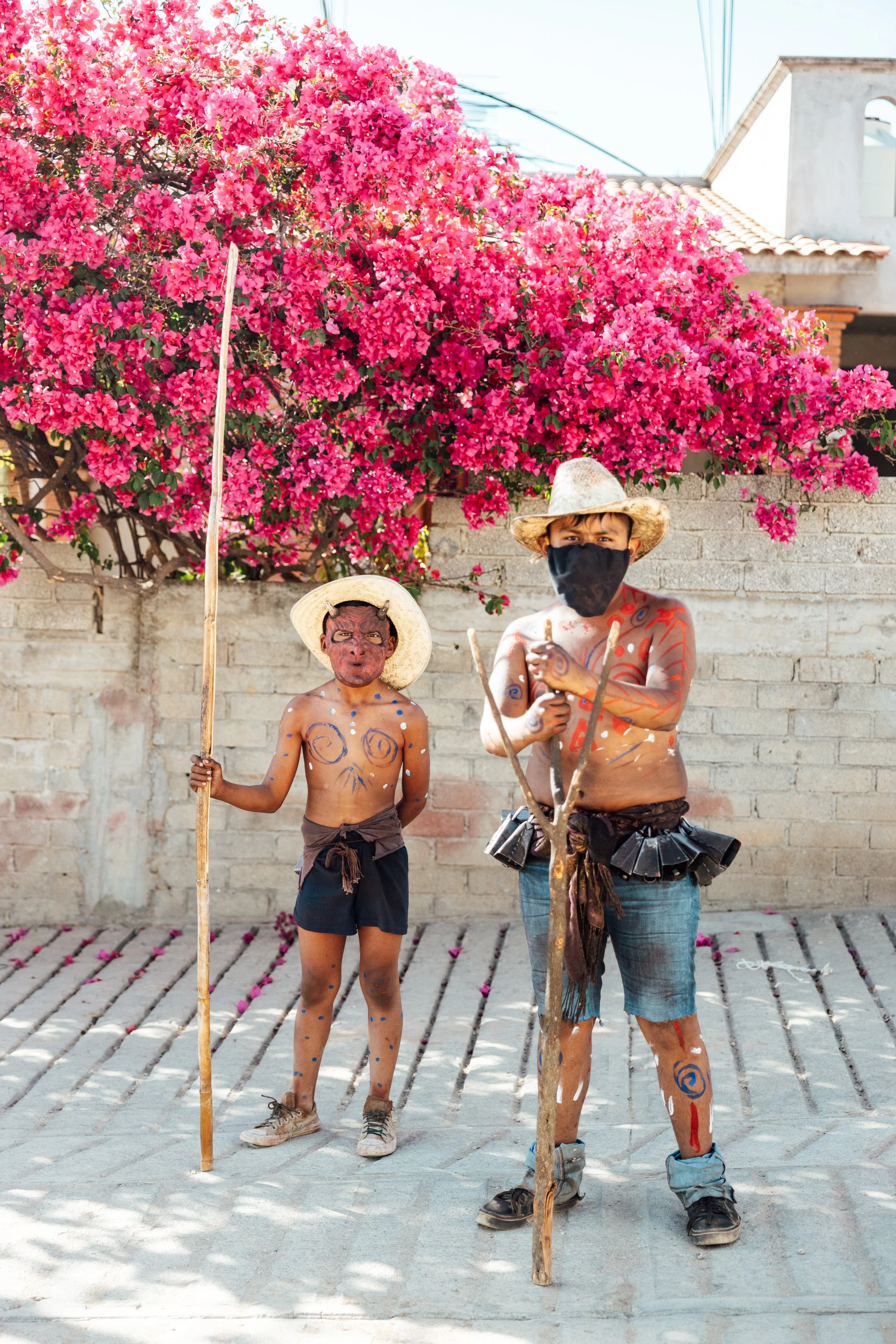 Two boys dressed as traditional Mexican dancers with painted faces, wearing hats, holding sticks, standing in front of a large pink bougainvillea bush and a brick wall.