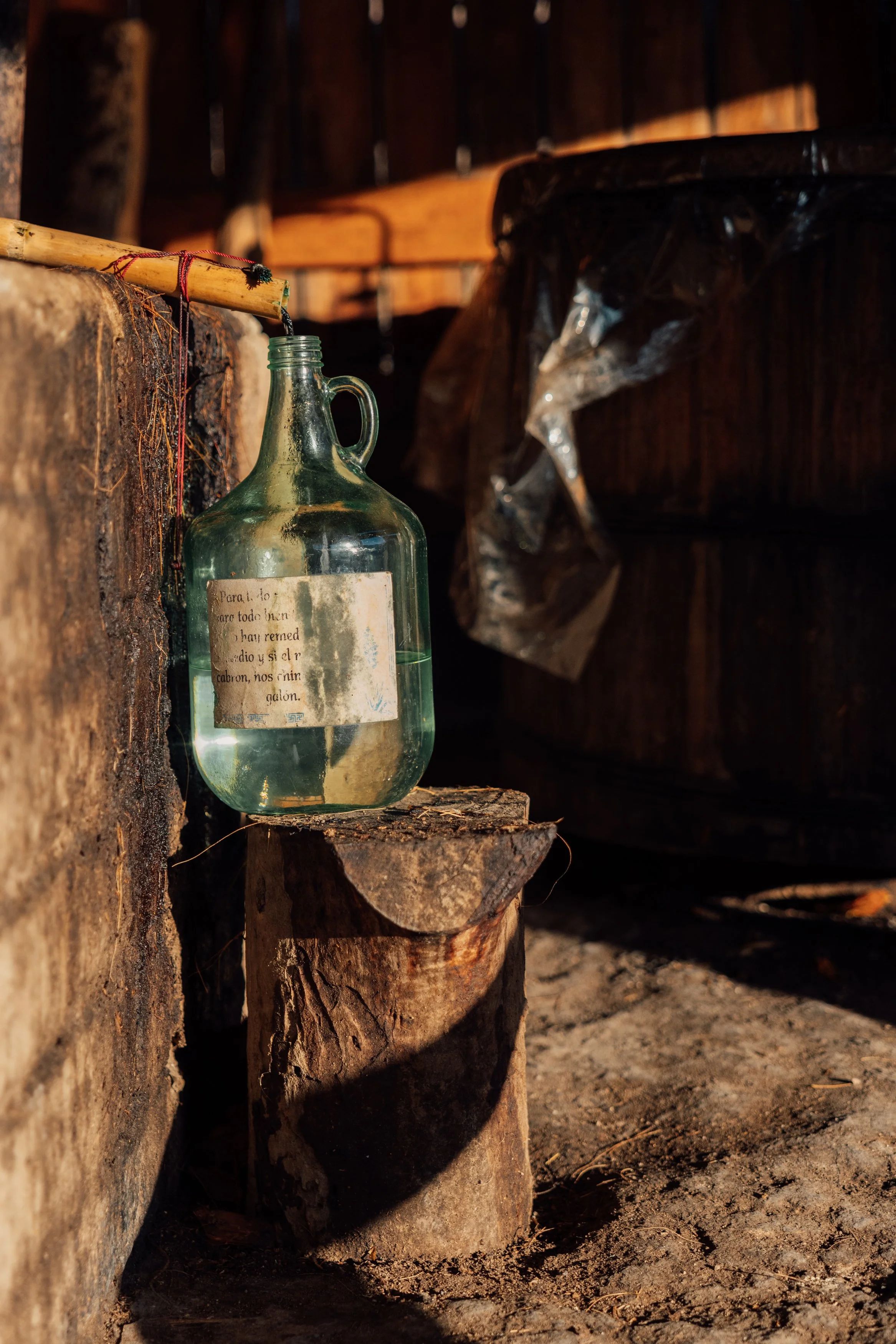 A glass jug with a paper label attached, placed on a cut piece of wood, with sunlight casting shadows on the ground and wooden wall in the background.