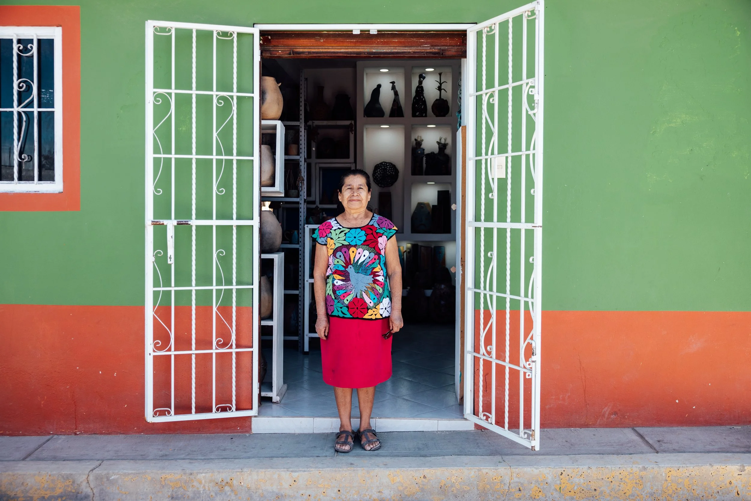 A woman standing in front of a green and orange building with open white metal gates, inside a store with pottery and art displays.