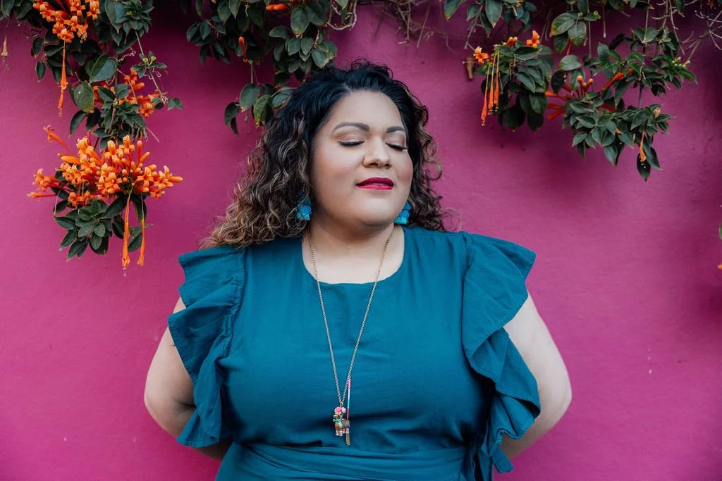 A woman with curly hair and blue earrings standing against a pink wall with hanging orange flowers, wearing a blue dress with ruffled sleeves and a necklace.