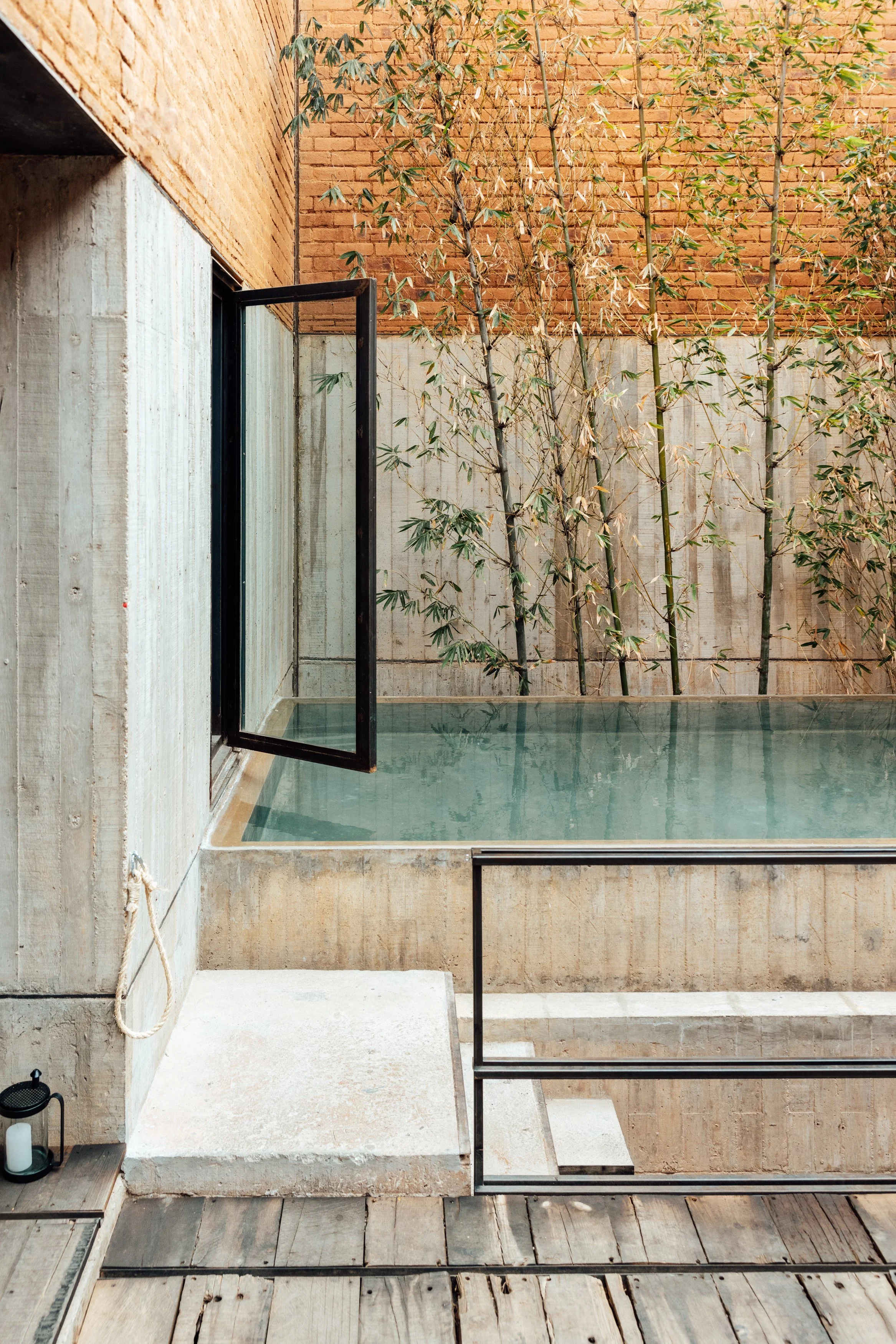 A small outdoor pool with clear water, surrounded by a wooden deck, a concrete step, and a black metal railing. There is a wall with wooden and brick sections, and tall bamboo plants growing behind the pool. An open window with a black frame is on th