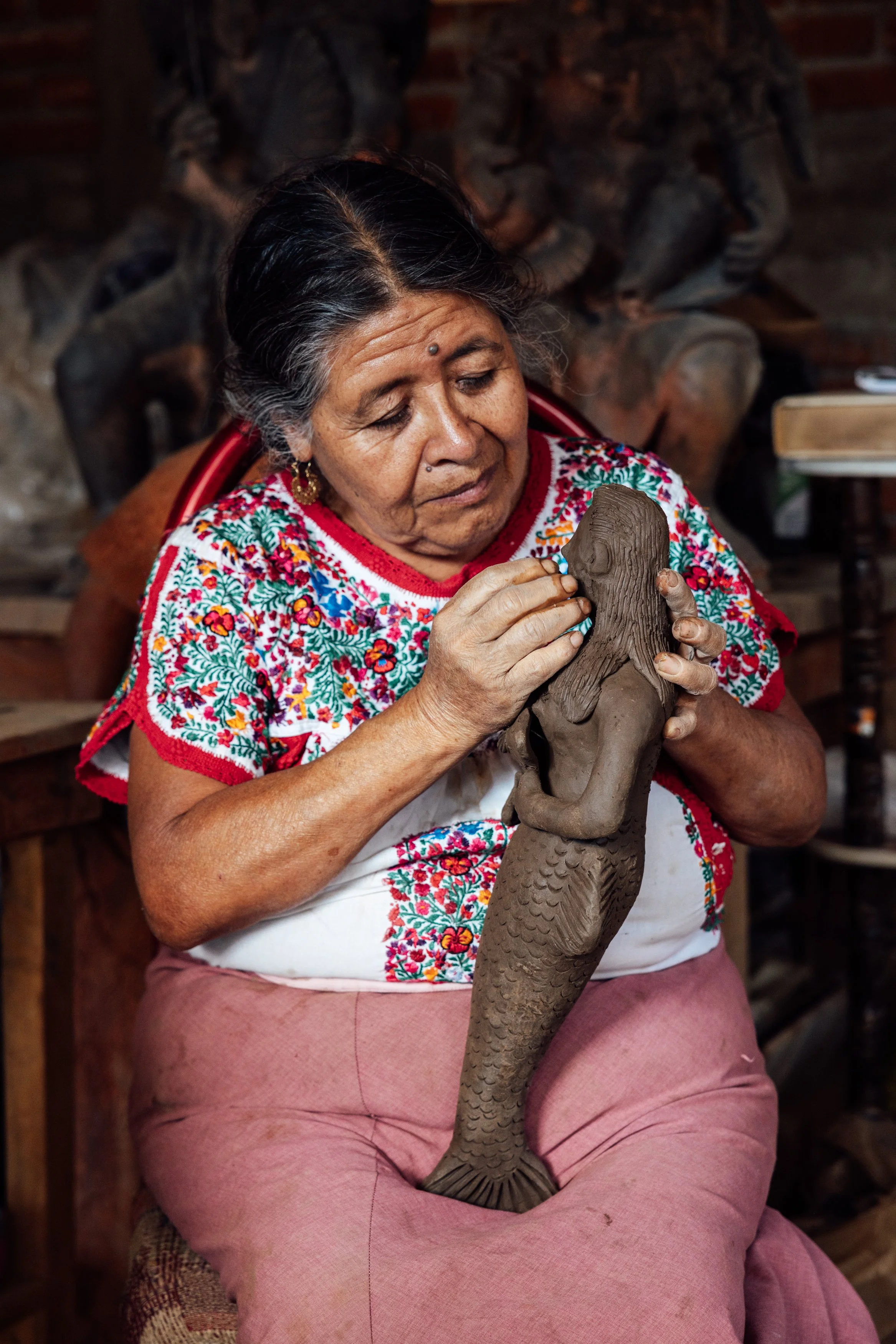 An elderly woman dressed in traditional colorful embroidered clothing is shaping a clay sculpture of a mermaid.