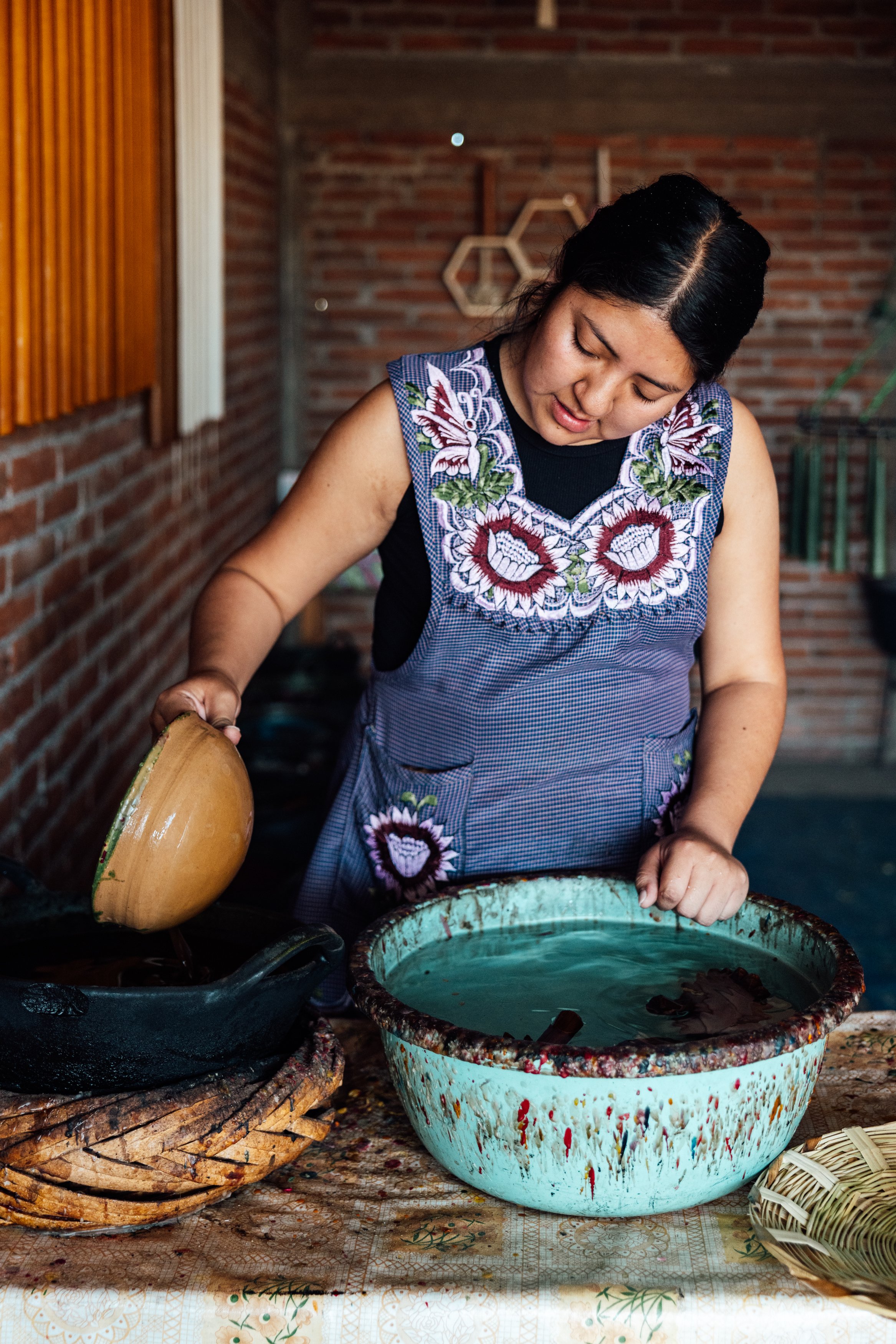 A woman in a decorative embroidered apron pouring liquid into a large ceramic basin, surrounded by pottery and a textured brick wall in the background.