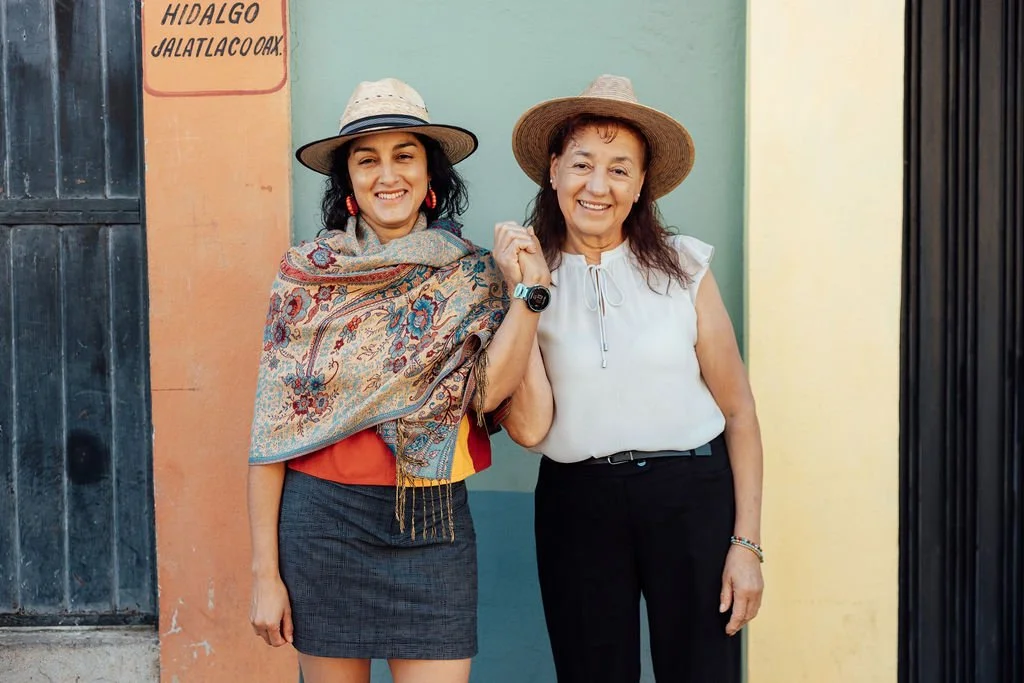 Two women smiling and holding hands, wearing straw hats, standing in front of a colorful wall.