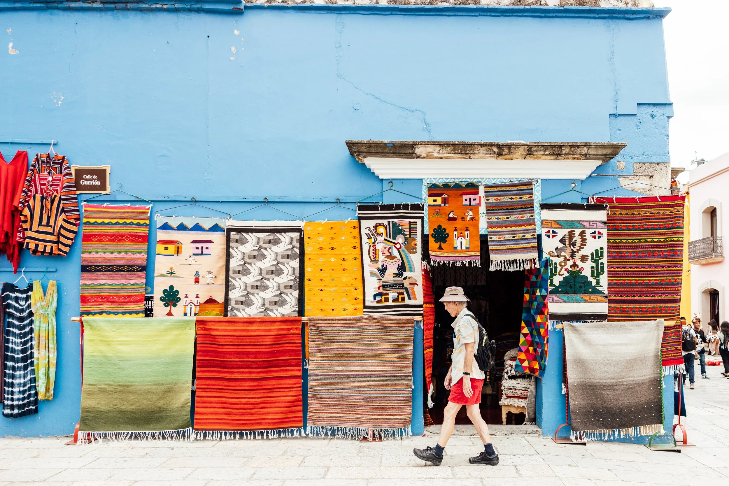 Colorful textiles and rugs displayed outside a shop with a bright blue wall, with a woman walking in front of it.