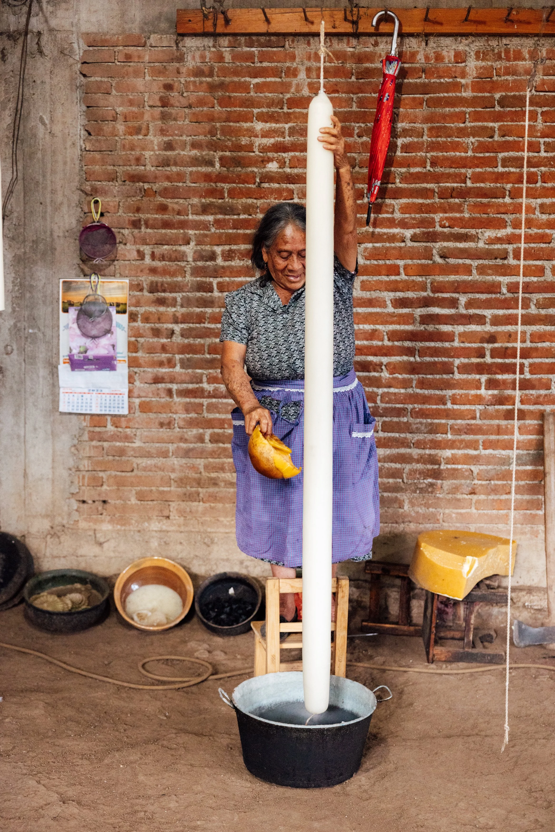 An elderly woman stands on a small wooden stool, holding a large white candle upright in a black bucket filled with water. She has a yellow object in her hand and is in front of a brick wall with various items hanging and placed around her, including