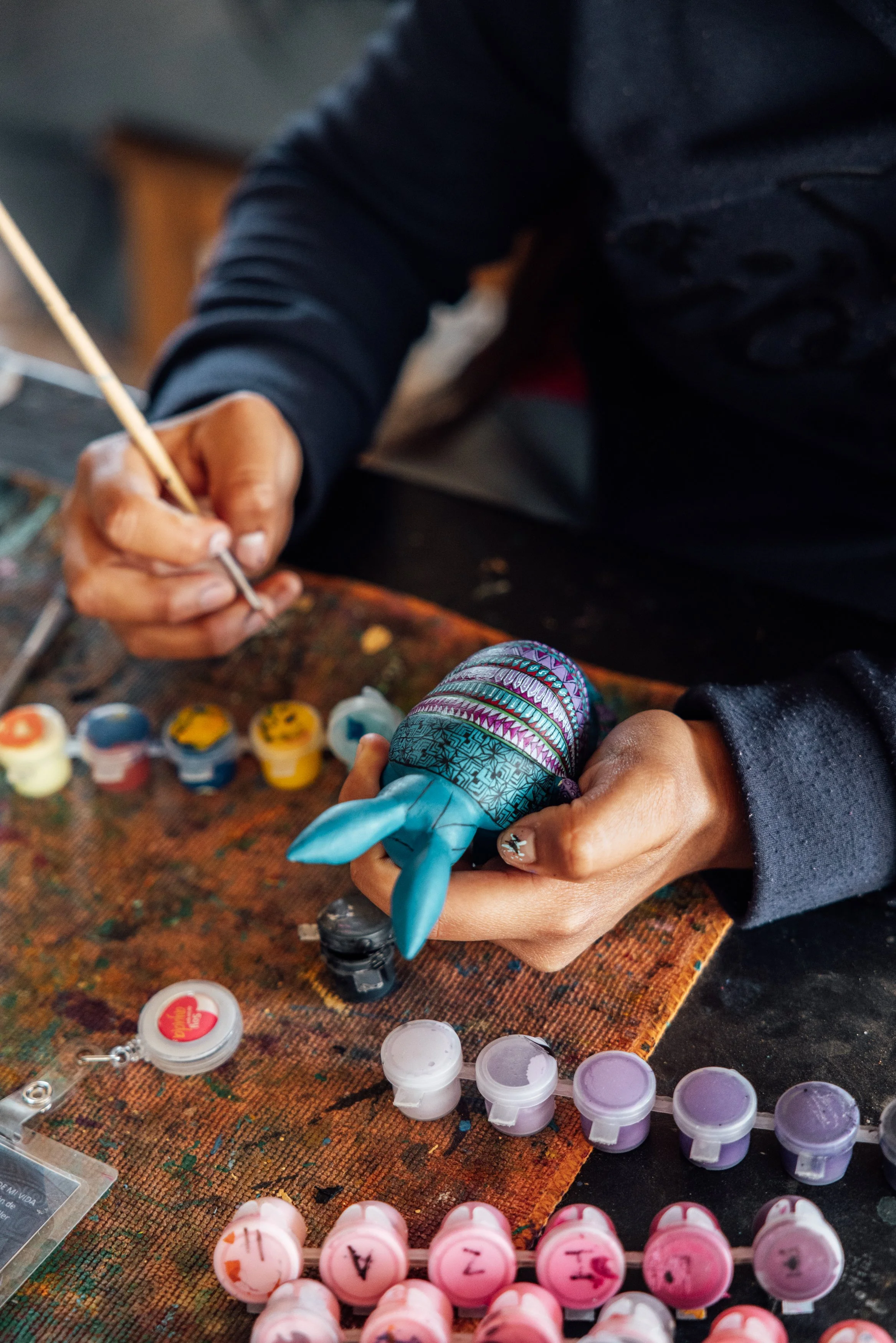 Person painting a small animal figurine with a paintbrush, surrounded by various small paint pots on a work surface.