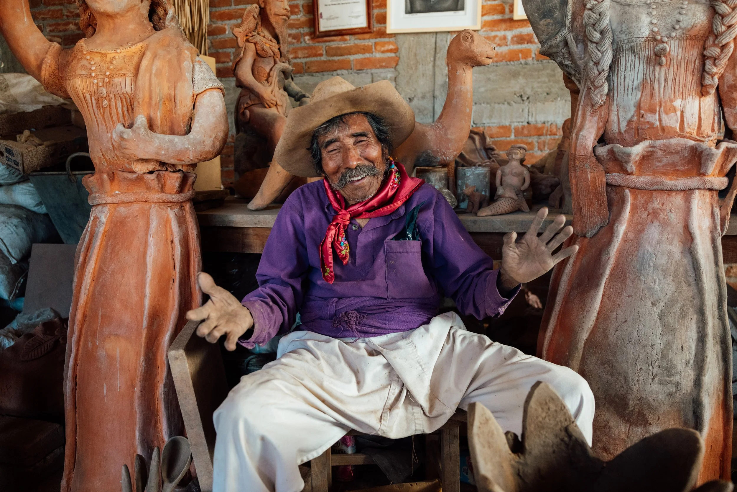 An elderly man with a mustache, wearing a cowboy hat, red neckerchief, purple shirt, and white pants, sitting and smiling in a workshop surrounded by large clay sculptures of animals and humanoid figures.