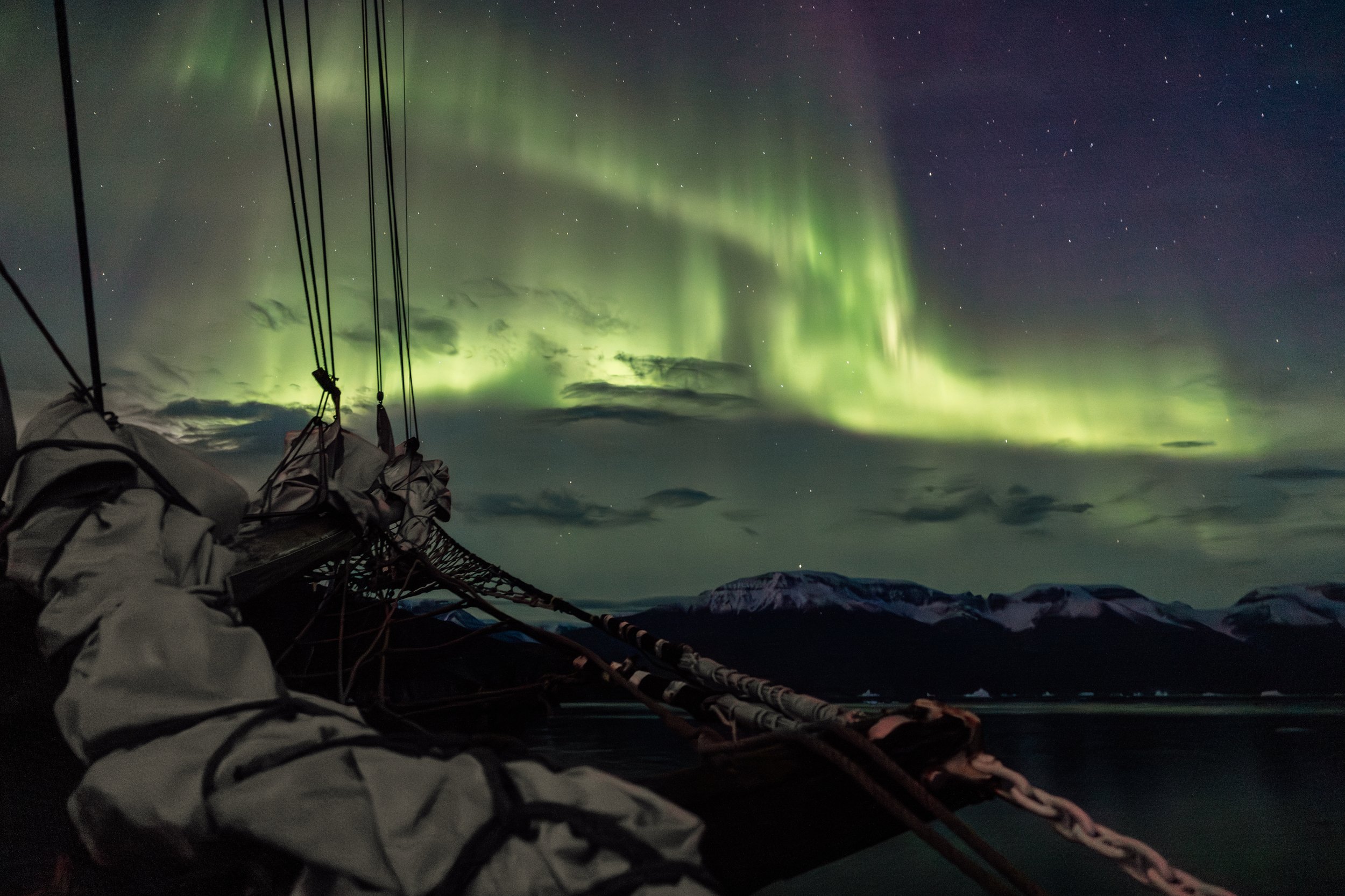 The photograph displays vibrant green and purple northern lights illuminating the night sky over a mountain range, with part of a boat or ship and its rigging visible in the foreground.