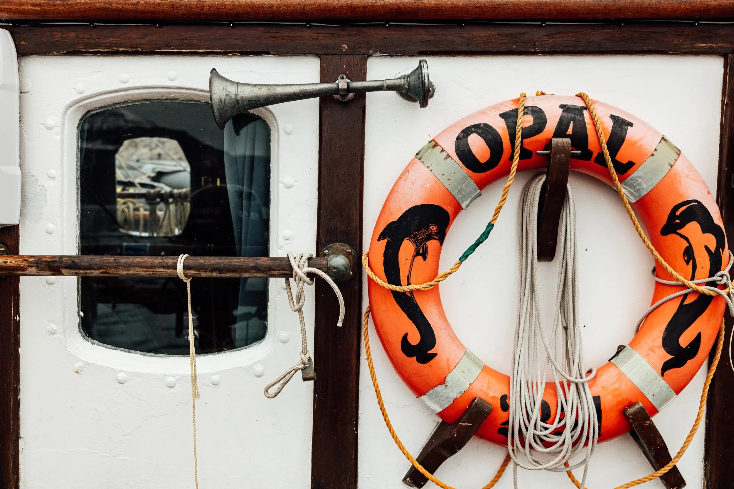 Close-up of a life preserver with the word 'OPEL' and dolphin illustrations hanging on a boat wall, next to a small window and a brass horn.