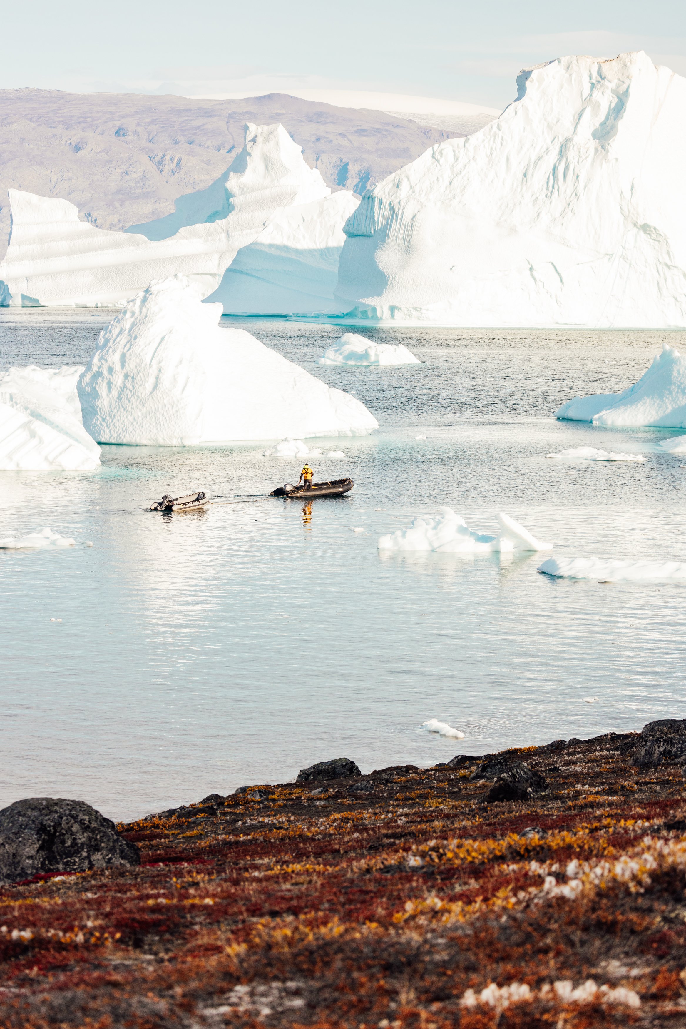 A person in a yellow jacket on a small inflatable boat near icebergs and large ice formations in a cold, Arctic-like landscape.