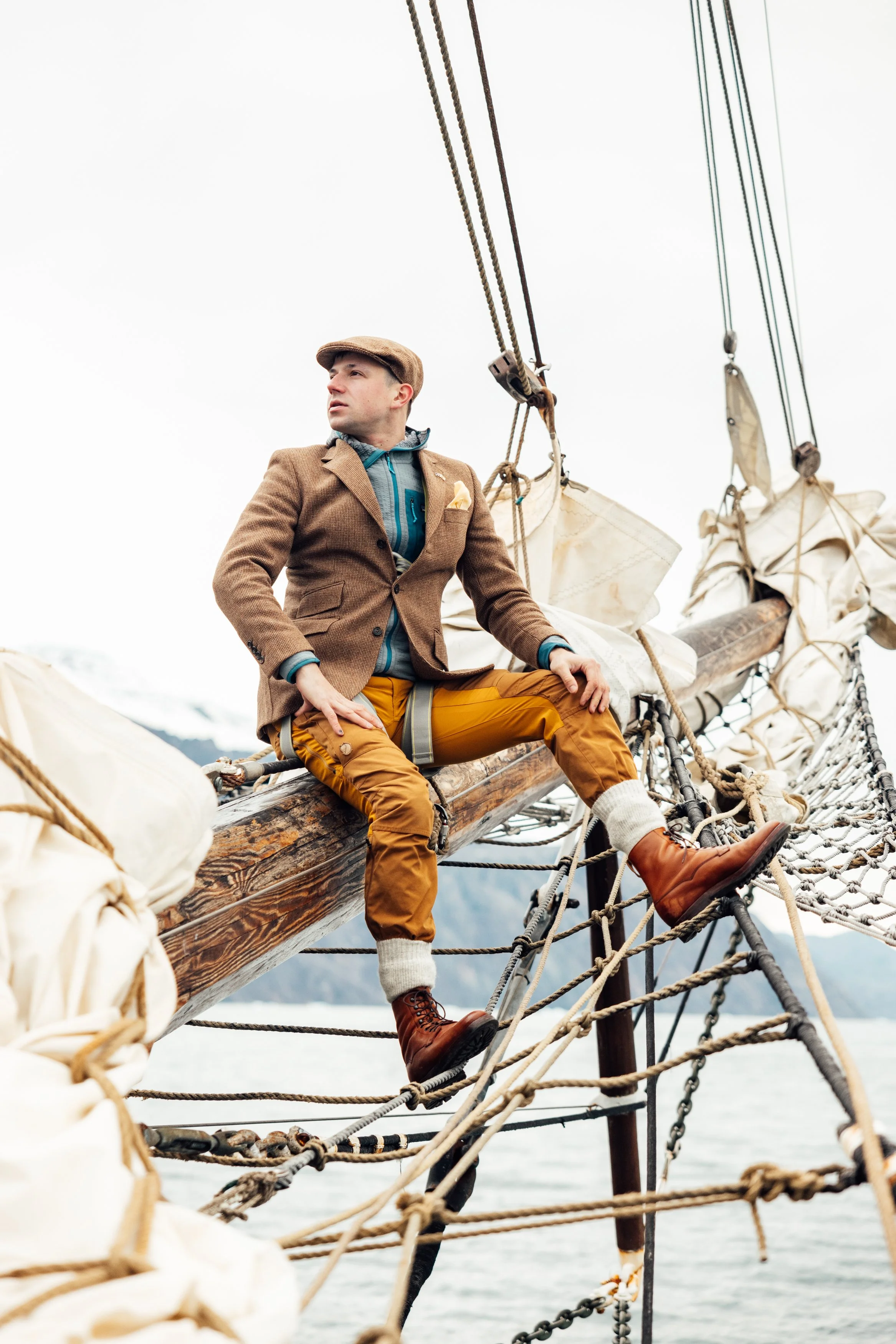 A man sitting on a sailing ship's mast with mountains and water in the background, wearing a brown jacket, yellow pants, and brown boots.
