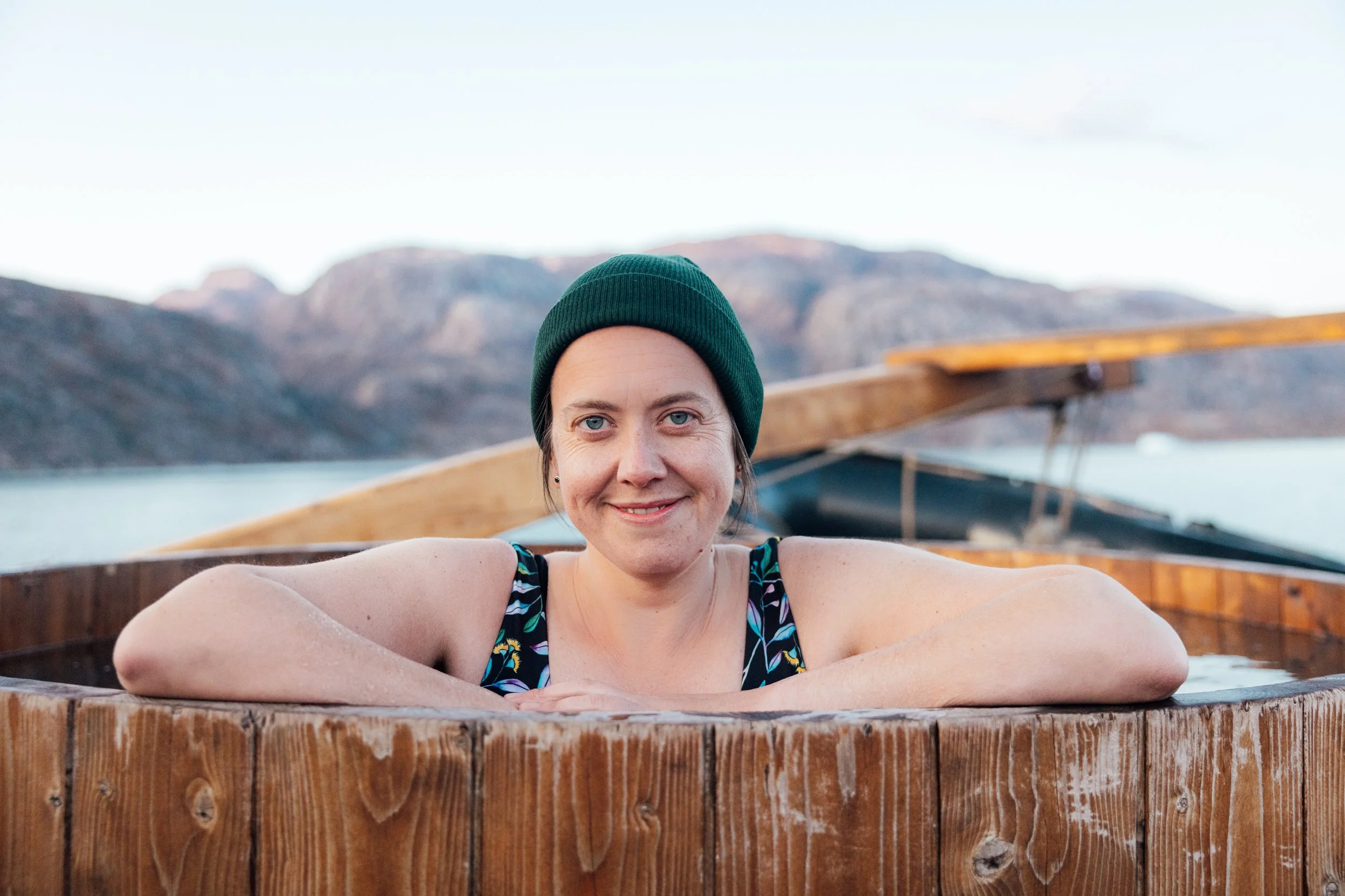 Woman with short hair, wearing a green beanie and a swimsuit, relaxing in a wooden hot tub outdoors with mountains and a lake in the background.