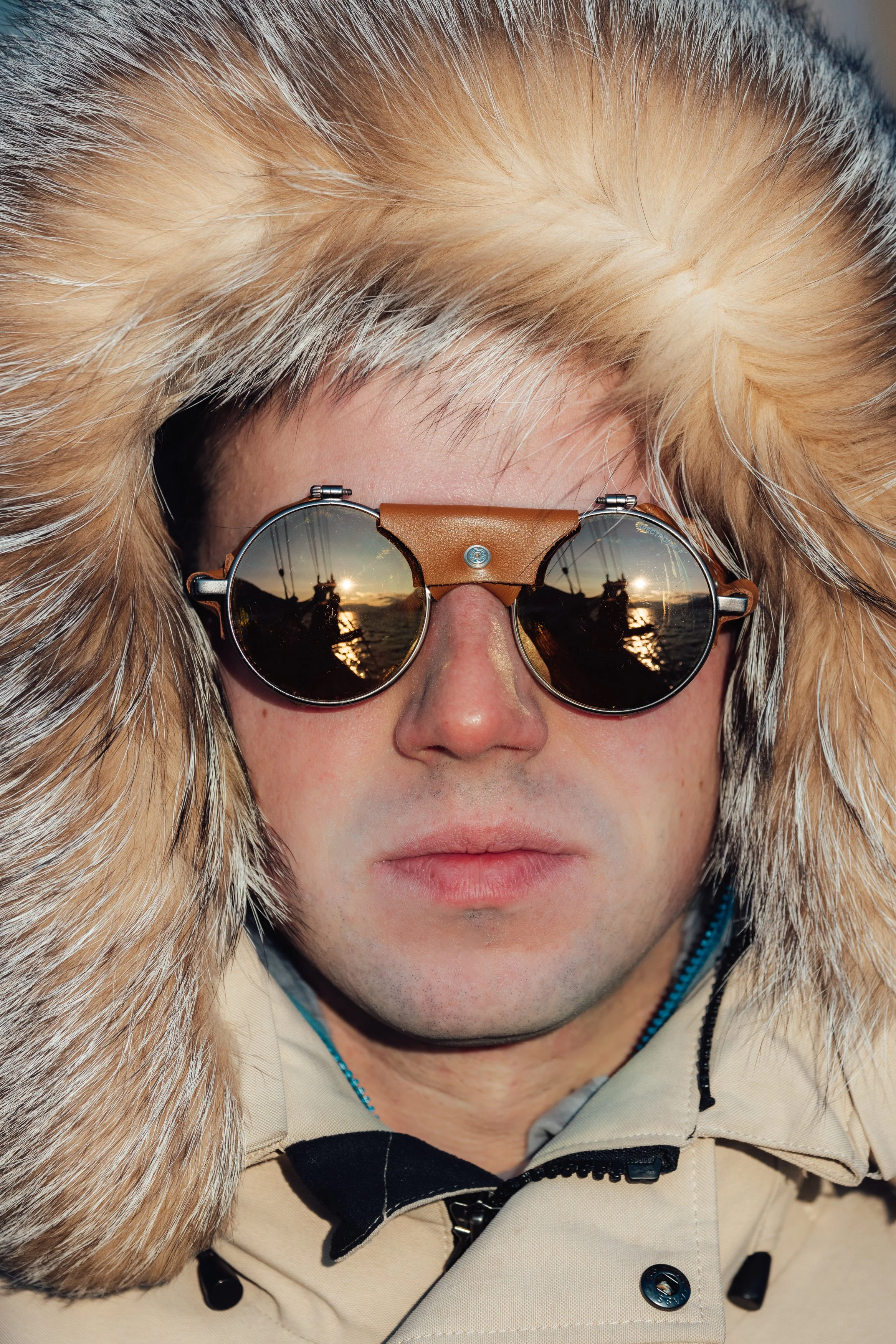 Close-up of a person wearing round sunglasses and a fur-lined hooded coat, with a reflection of a sunset and boats on the glasses, outdoors during sunset.