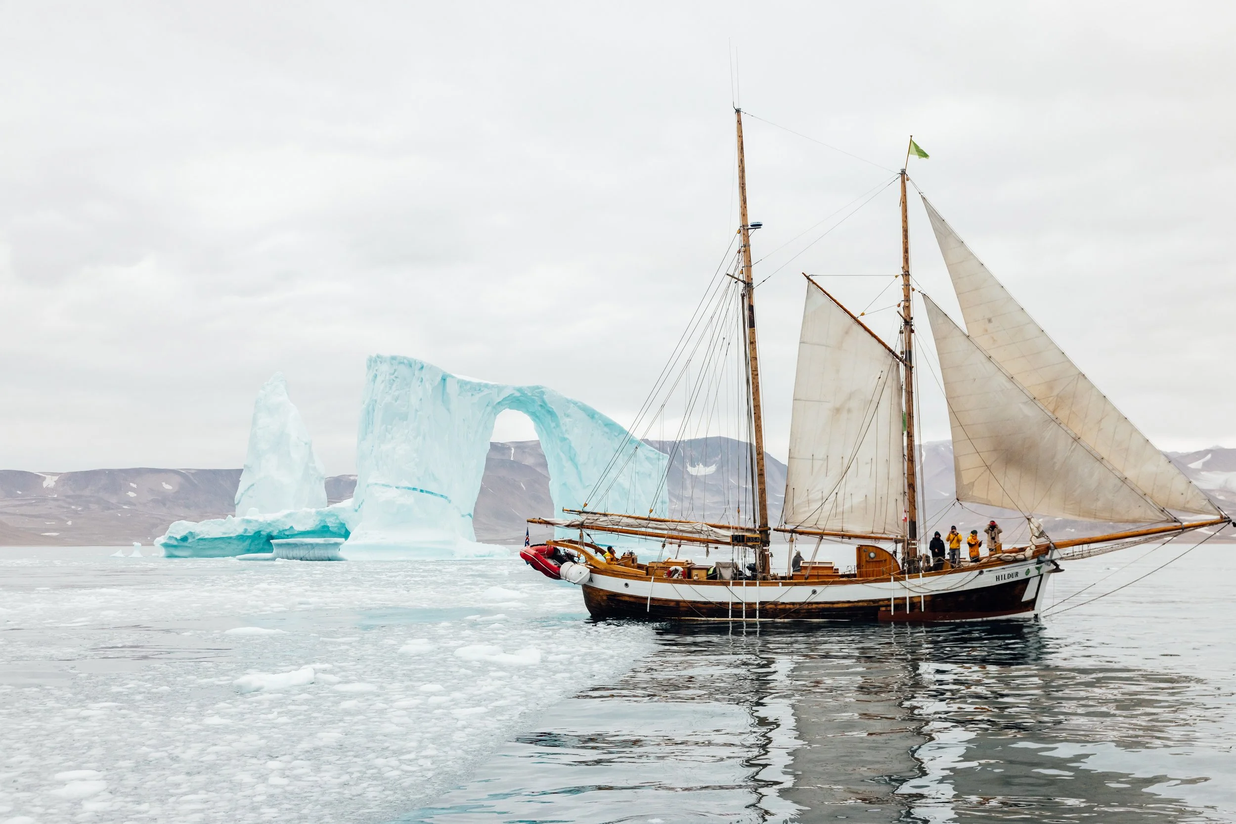 A wooden sailboat with white sails sailing near a large iceberg in icy waters, with mountains in the background.