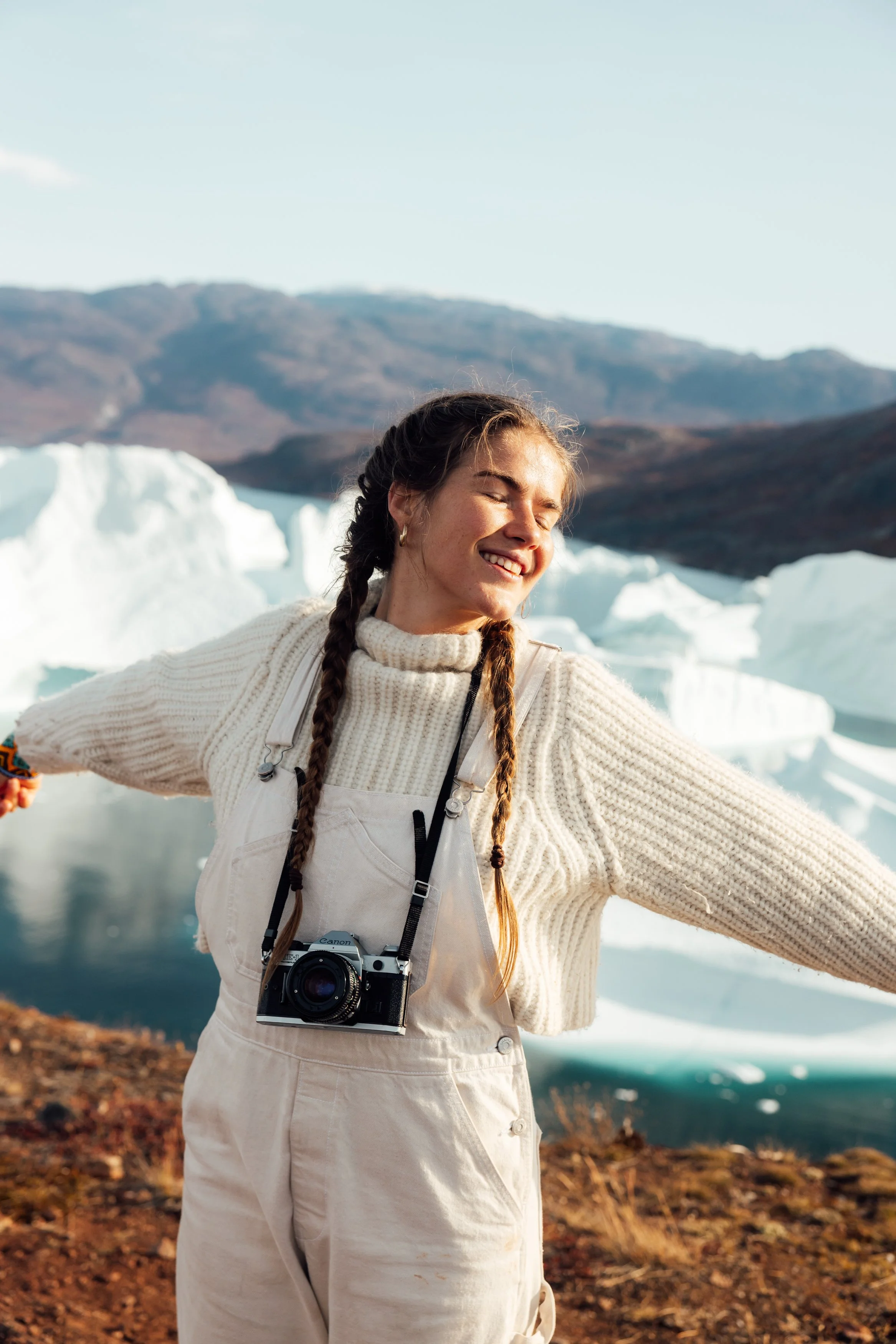 A woman with braided hair smiling with arms outstretched in front of icebergs and mountains, wearing a white sweater, overalls, and a camera around her neck.