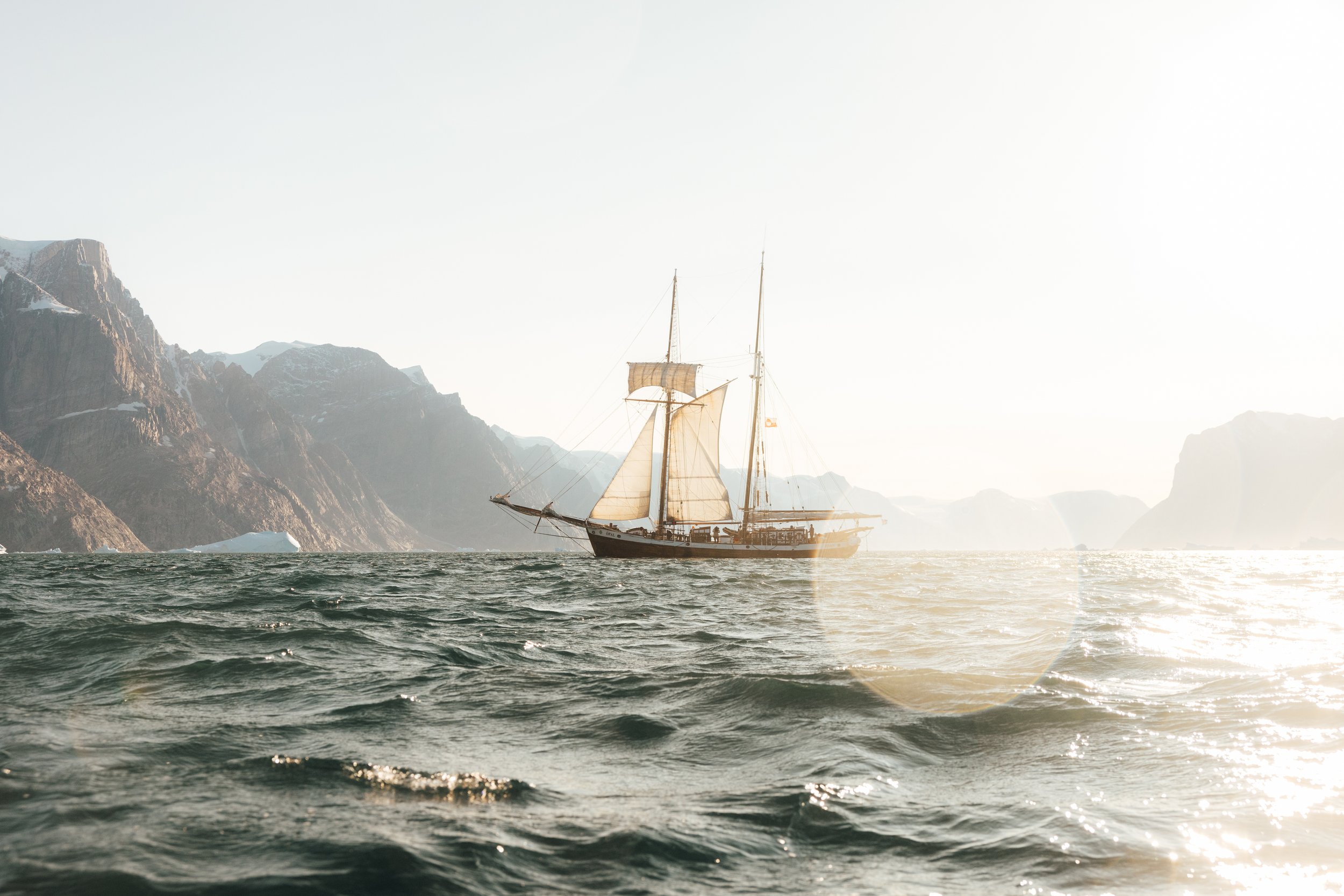 A sailing ship on the ocean with mountains in the background and sunlight reflecting on the water.