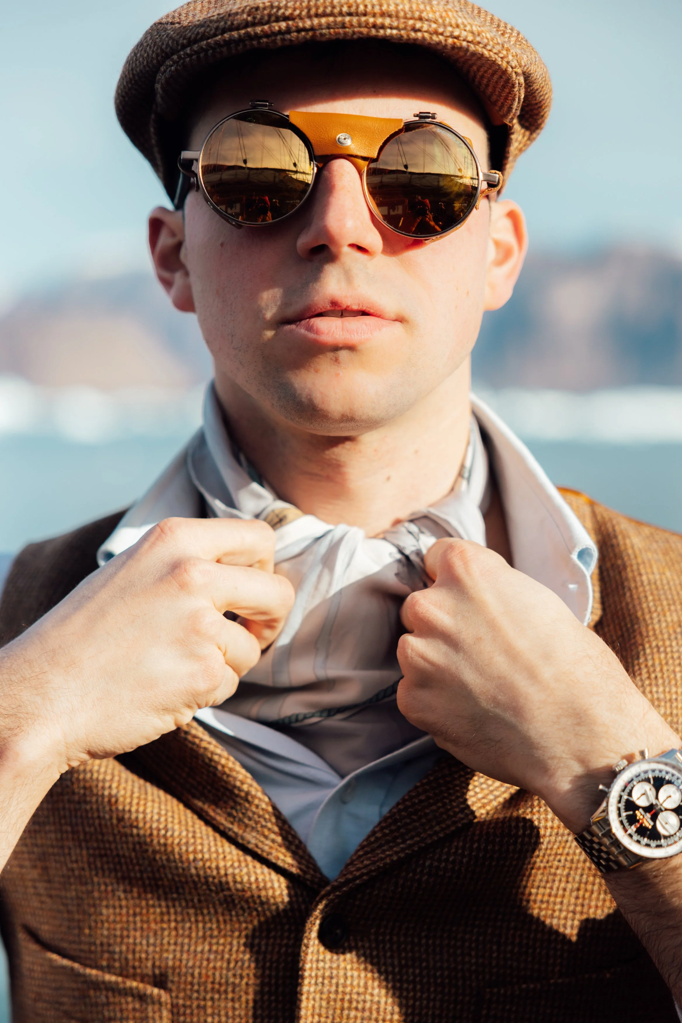 Close-up of a man wearing stylish sunglasses, a brown  textured jacket, adjusting a light-colored scarf around his neck, with a blurred outdoor background.
