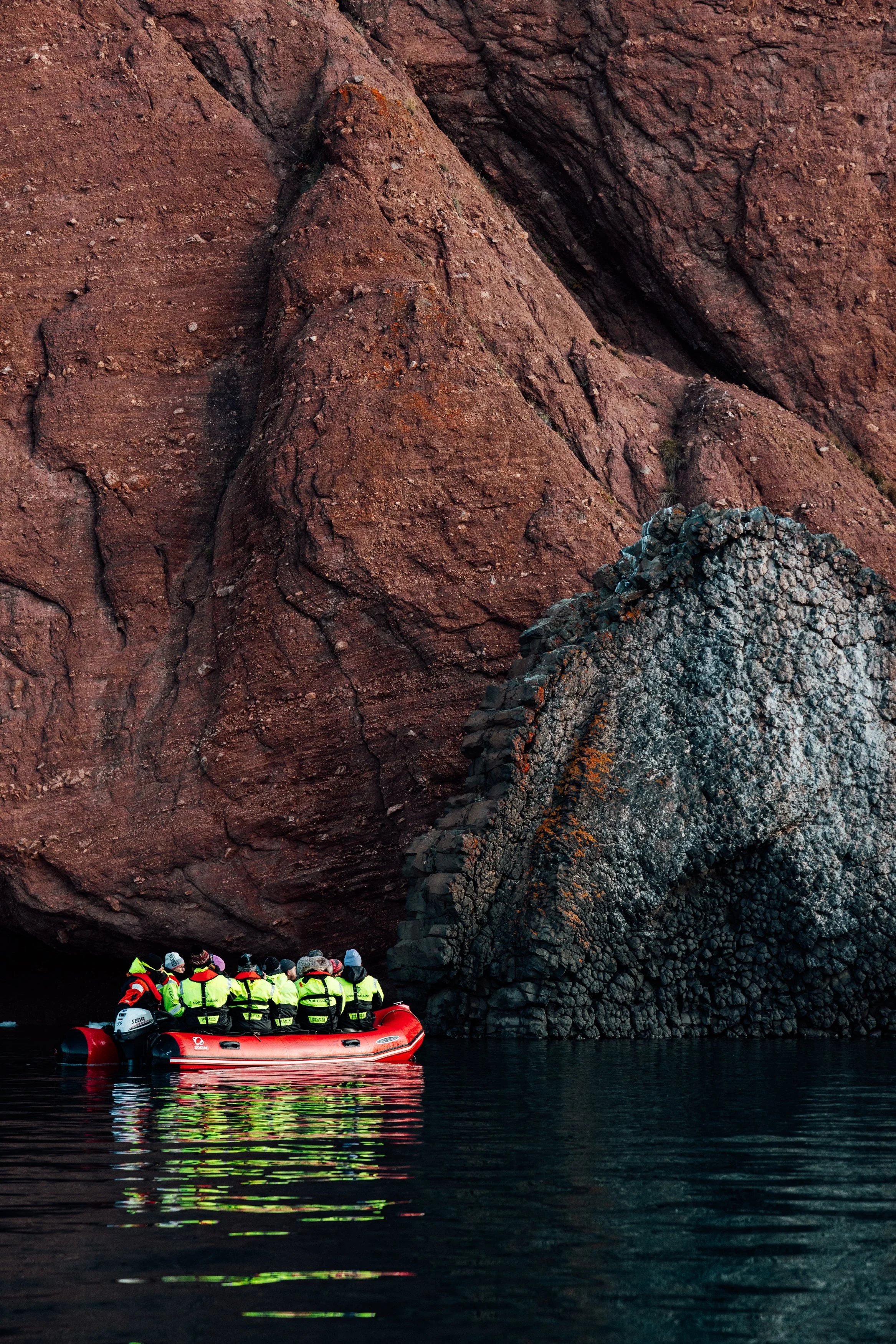 A group of people wearing life jackets and warm clothing on a red inflatable boat in front of large, reddish-brown rocky cliffs.