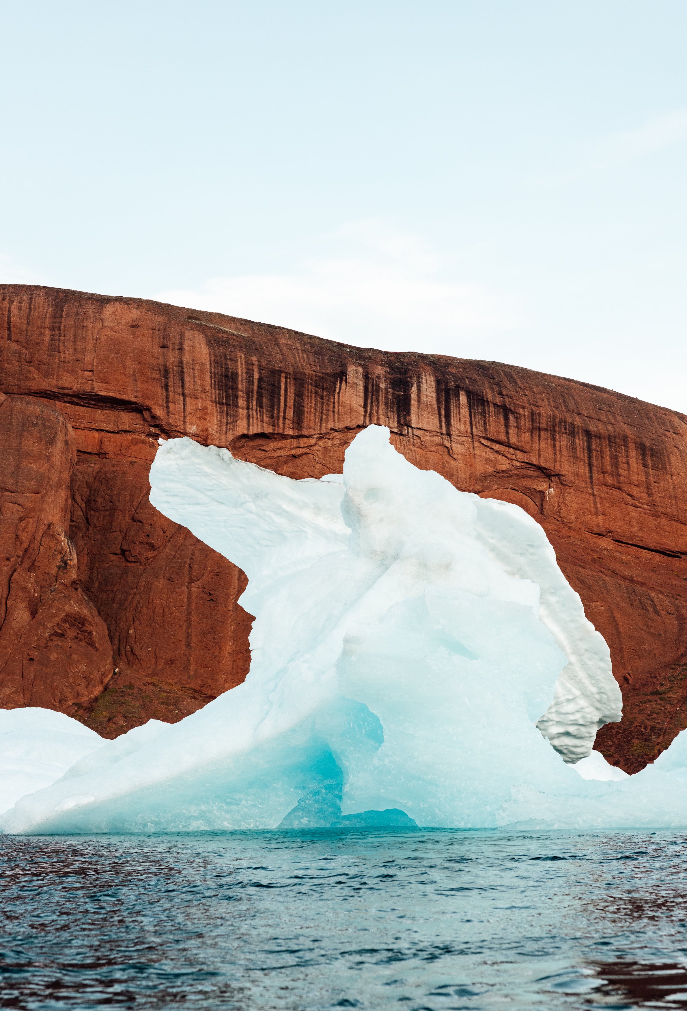 Iceberg in the shape of an animal with a red rock formation in the background, floating on water.