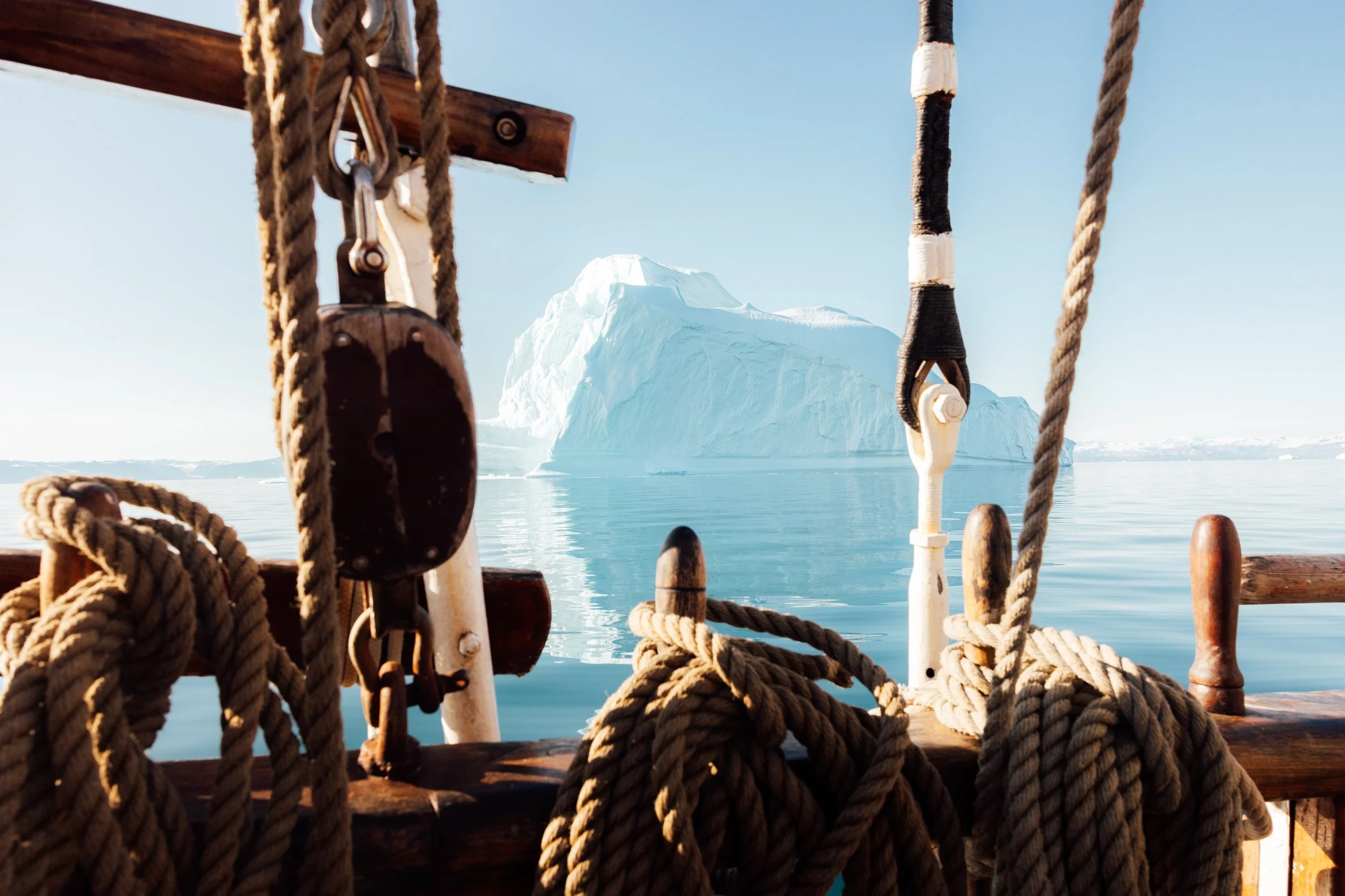 View from a boat showing ropes and pulleys in foreground, with a large iceberg in the distance on a calm sea under a clear sky.